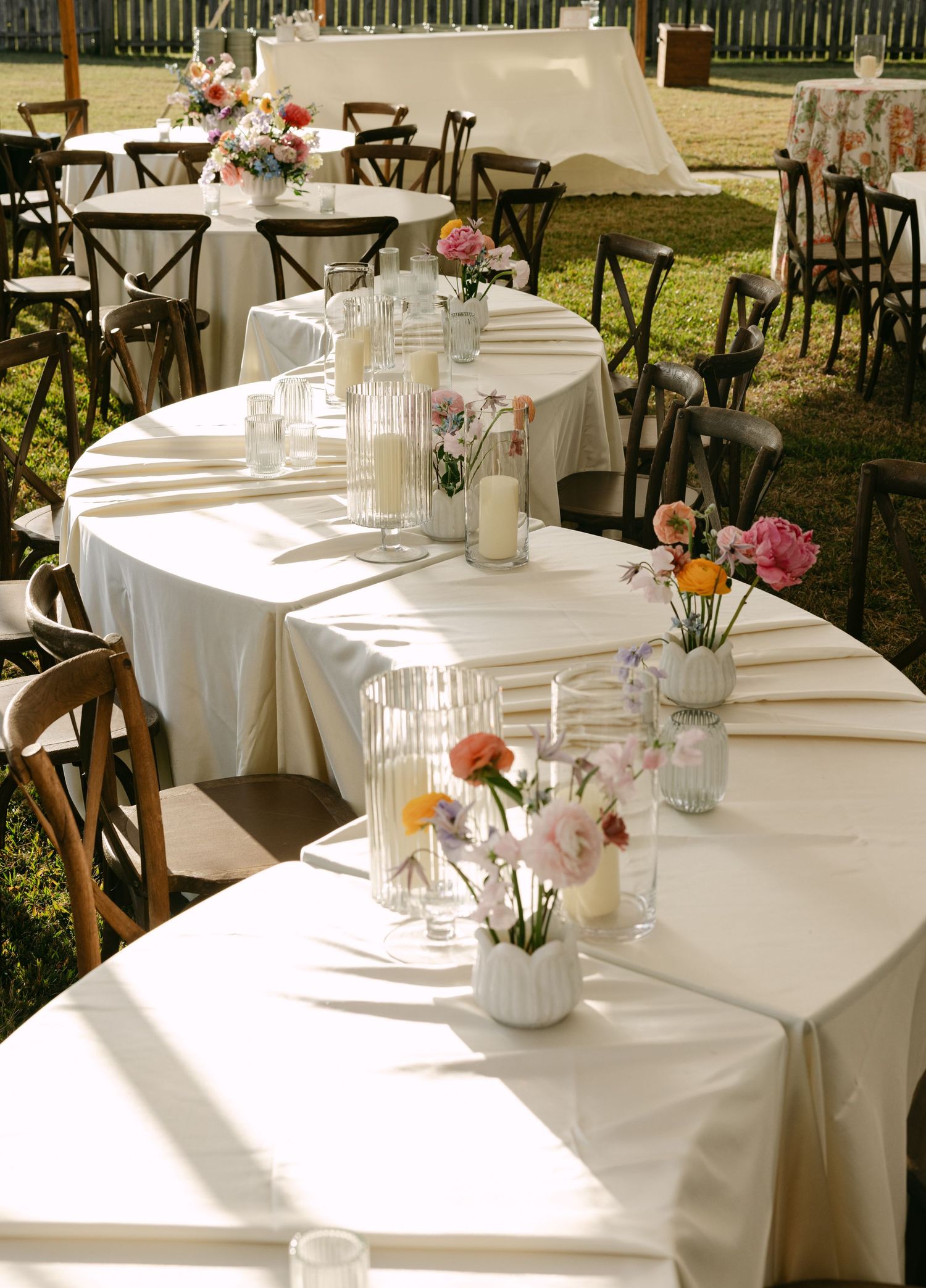 White linen-covered tables with rustic wooden chairs and small floral centerpieces set up for an outdoor wedding reception.