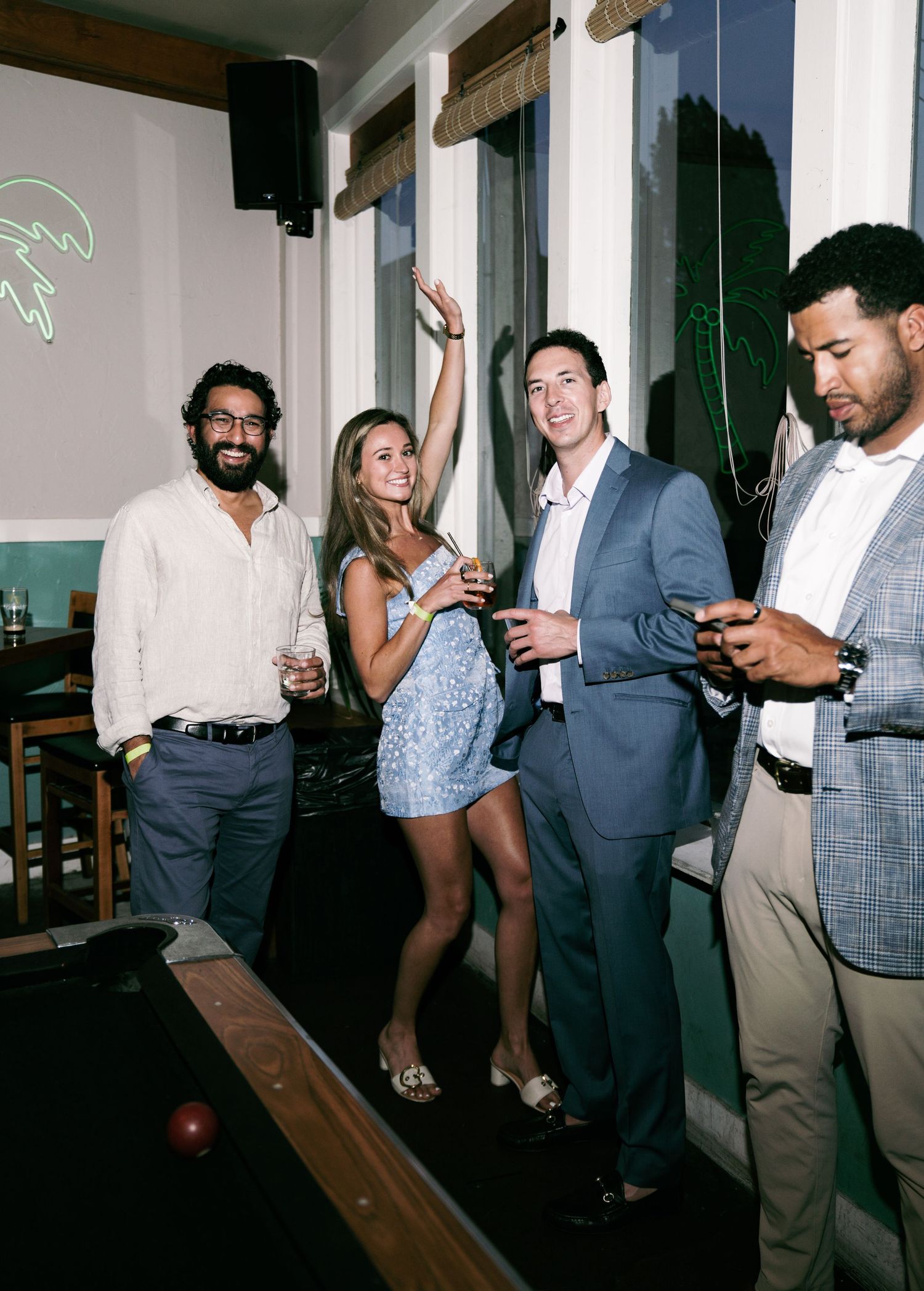 Four young adults socializing at an indoor party near a pool table, woman dancing with raised arm.