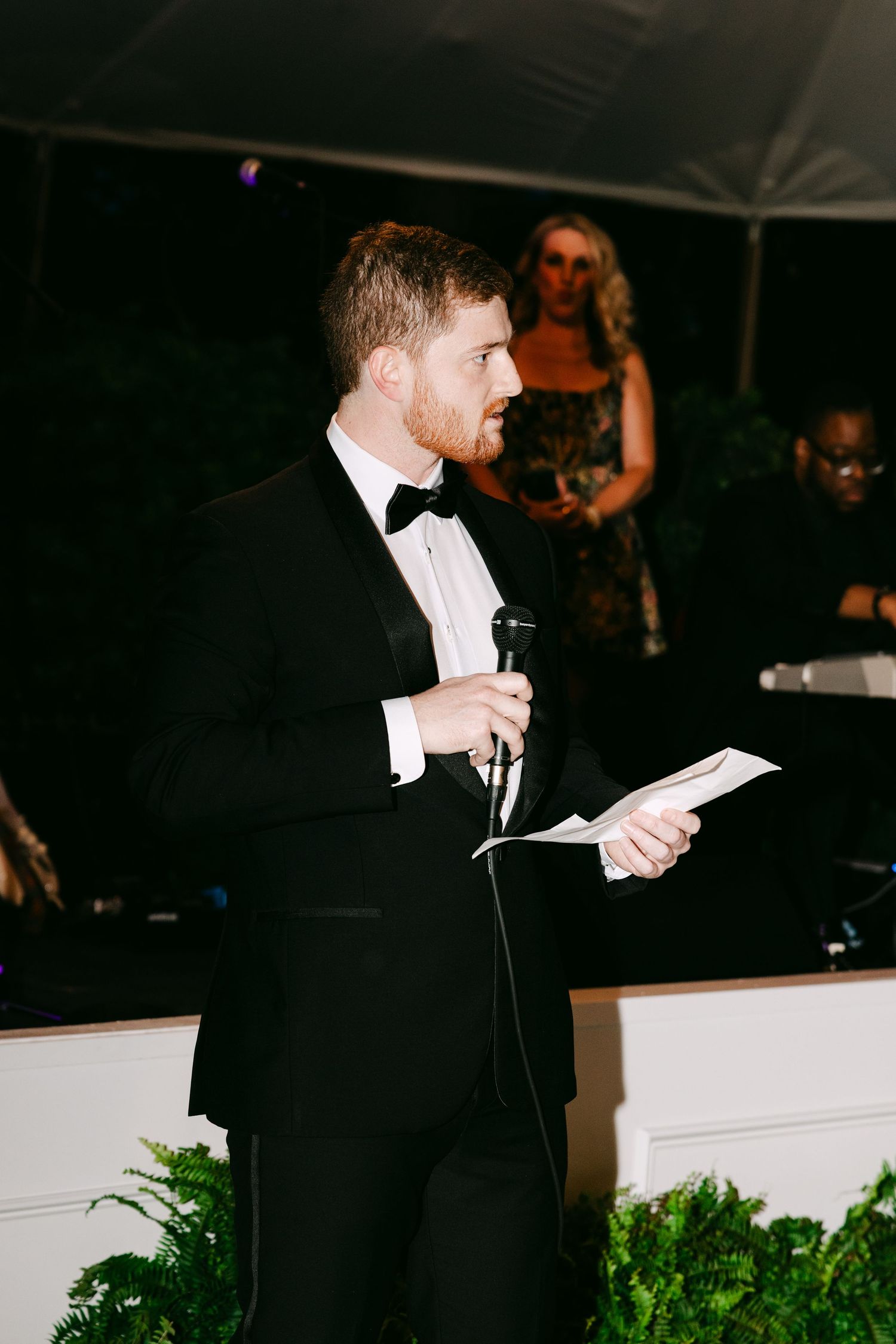 A man in a tuxedo delivers a wedding speech while holding a microphone and notes at an elegant evening reception.