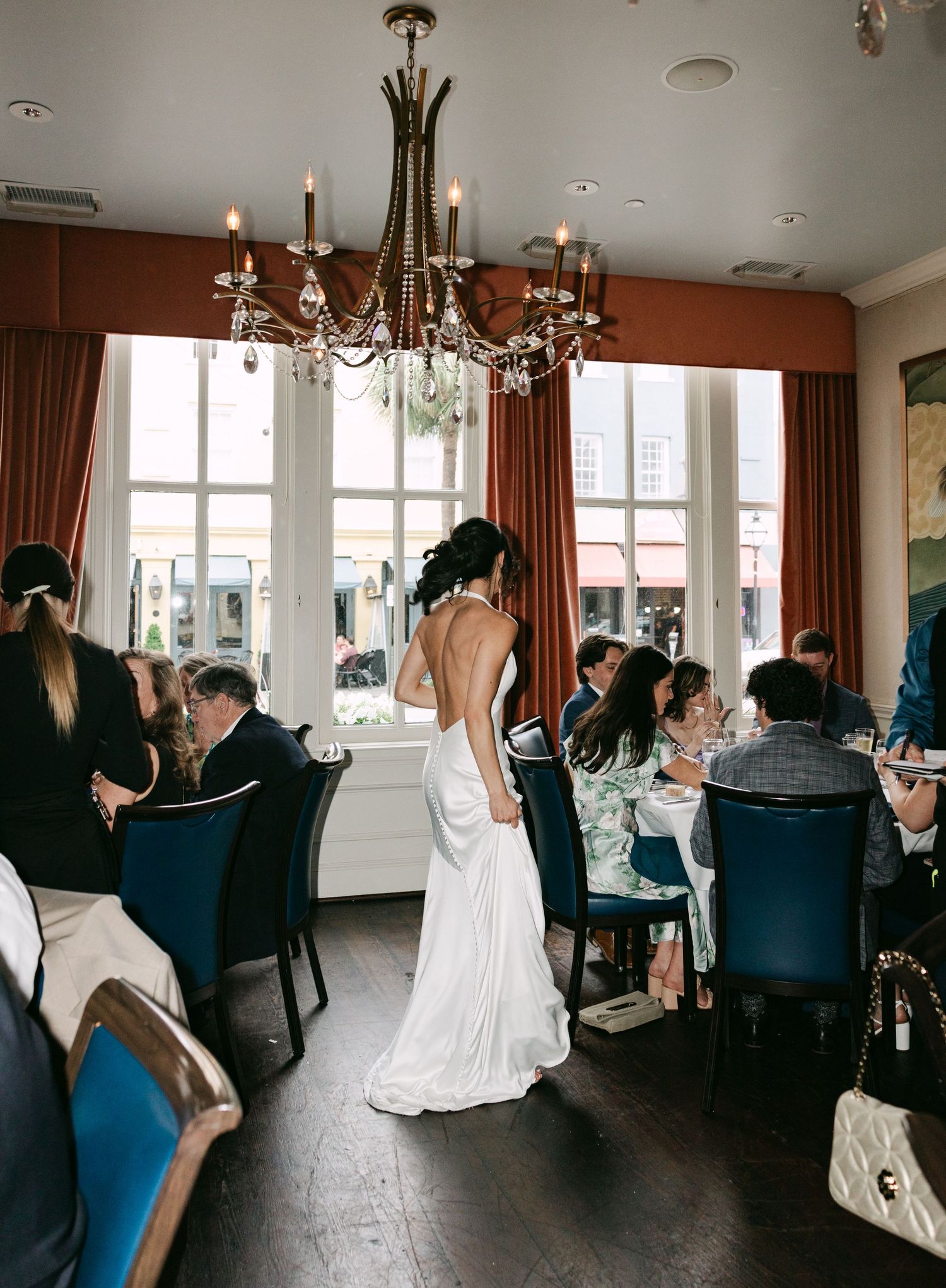 Bride in white gown stands among seated wedding guests in elegant restaurant with chandelier and large windows.