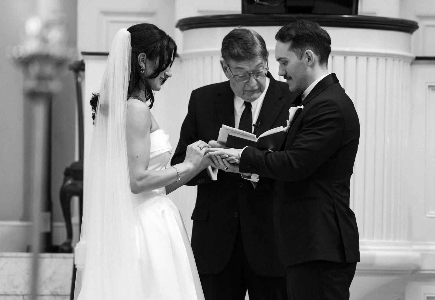 Black and white photo of a bride and groom exchanging rings during a wedding ceremony with an officiant.