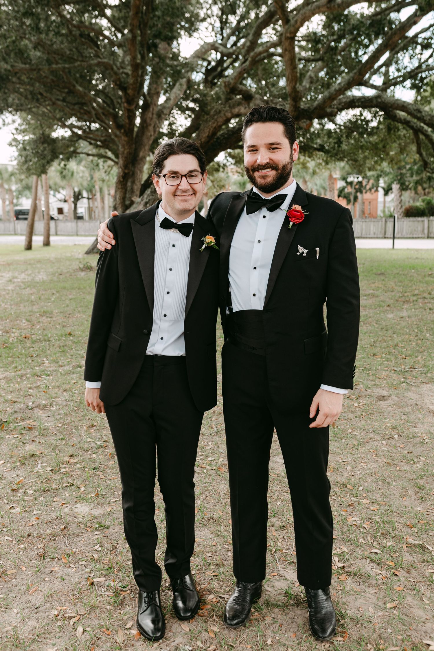 Two men in black tuxedos with bow ties posing outdoors under large oak trees.