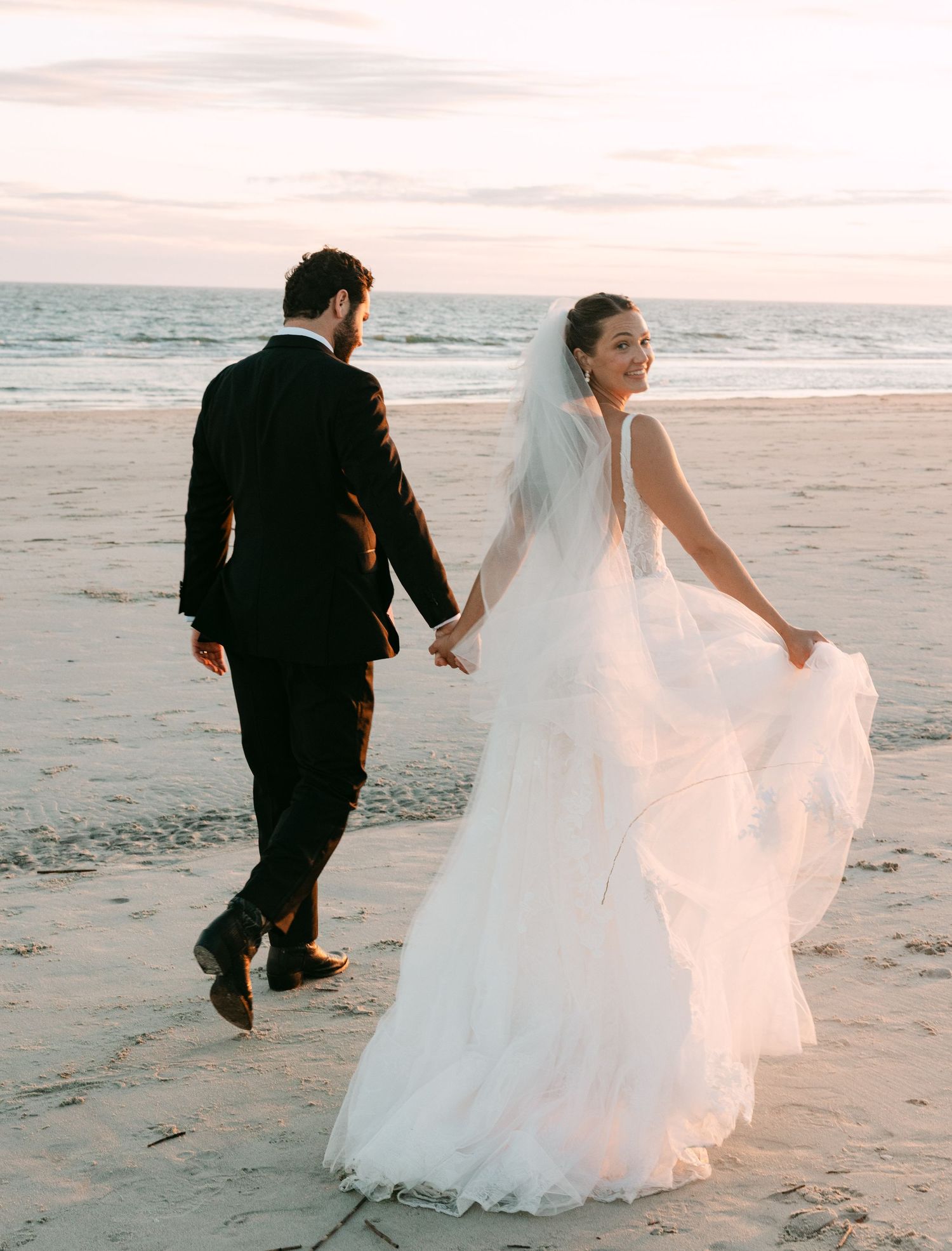 Bride in flowing lace gown and groom in black tuxedo walk hand-in-hand on beach at sunset.