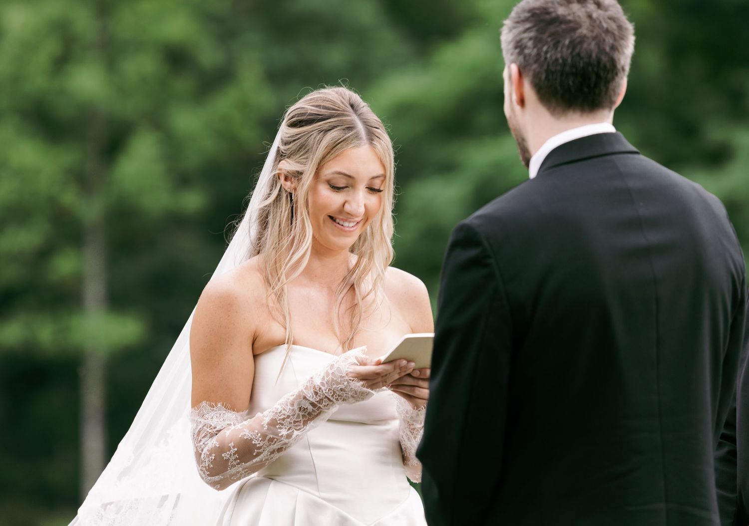 Bride in white gown and veil reads wedding vows while smiling, standing outdoors with groom in black suit.