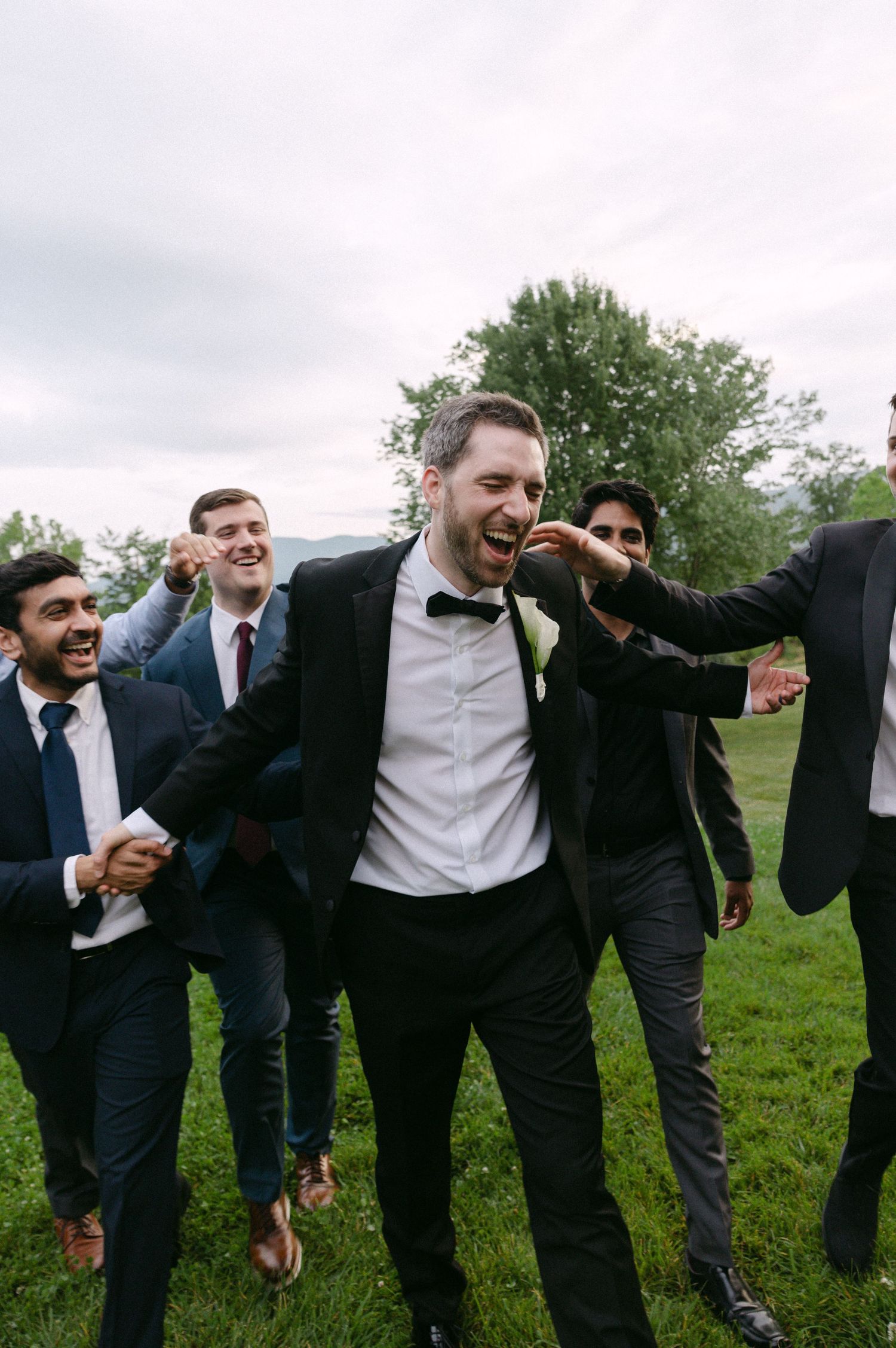 Groom laughing joyfully with groomsmen in suits on a green lawn with overcast sky backdrop.
