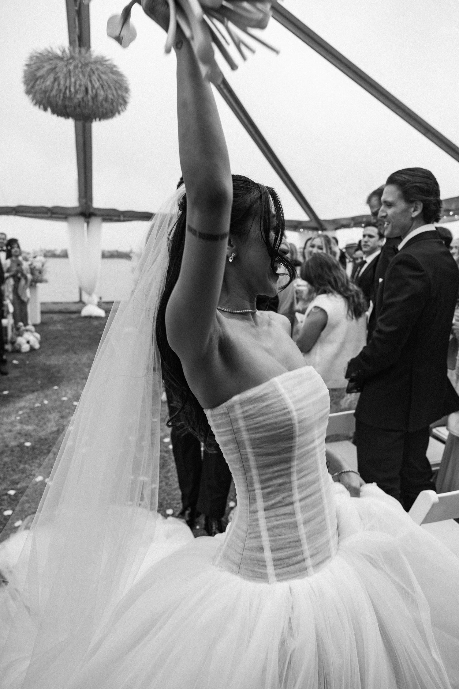 Black and white photo of a bride in a strapless ballgown raising her bouquet in celebration at her wedding ceremony.
