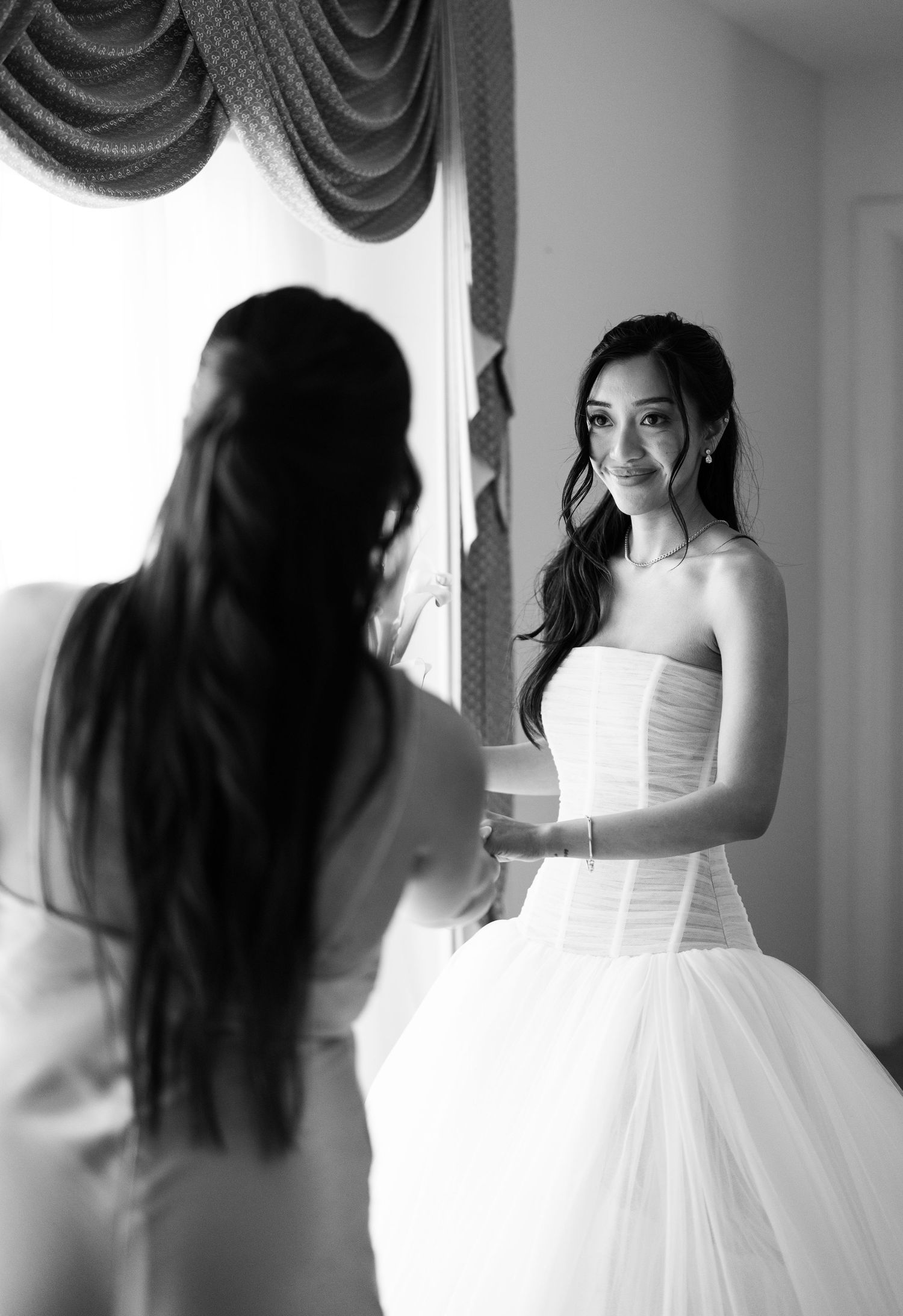 Bride in strapless white wedding gown smiles at reflection in mirror in black and white bridal preparation photo.