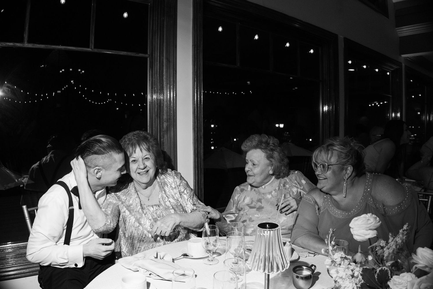 Black and white photo of elderly women laughing at a wedding reception dinner table with string lights in background.