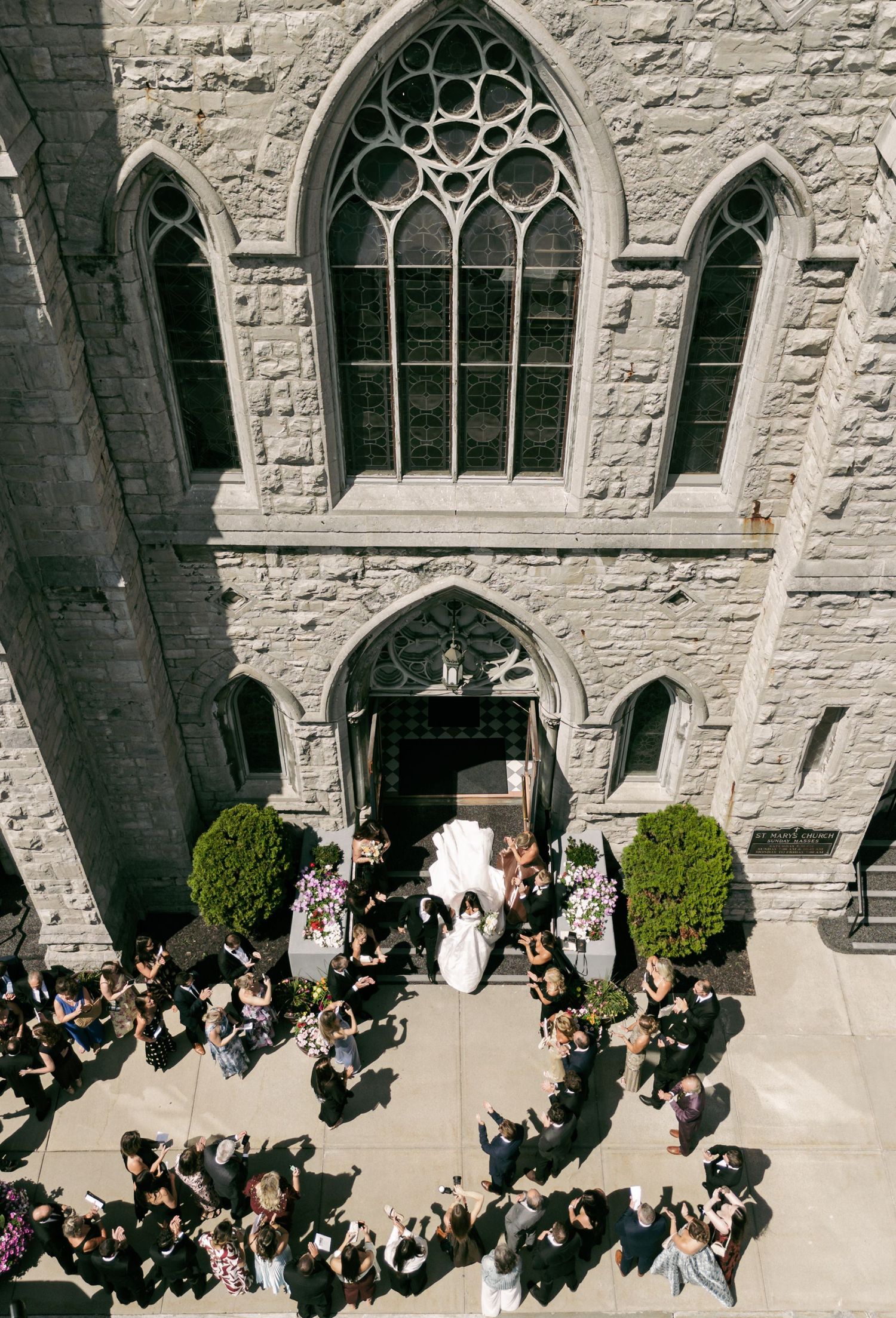 Aerial view of bride exiting gothic stone church surrounded by wedding guests on sunny day.