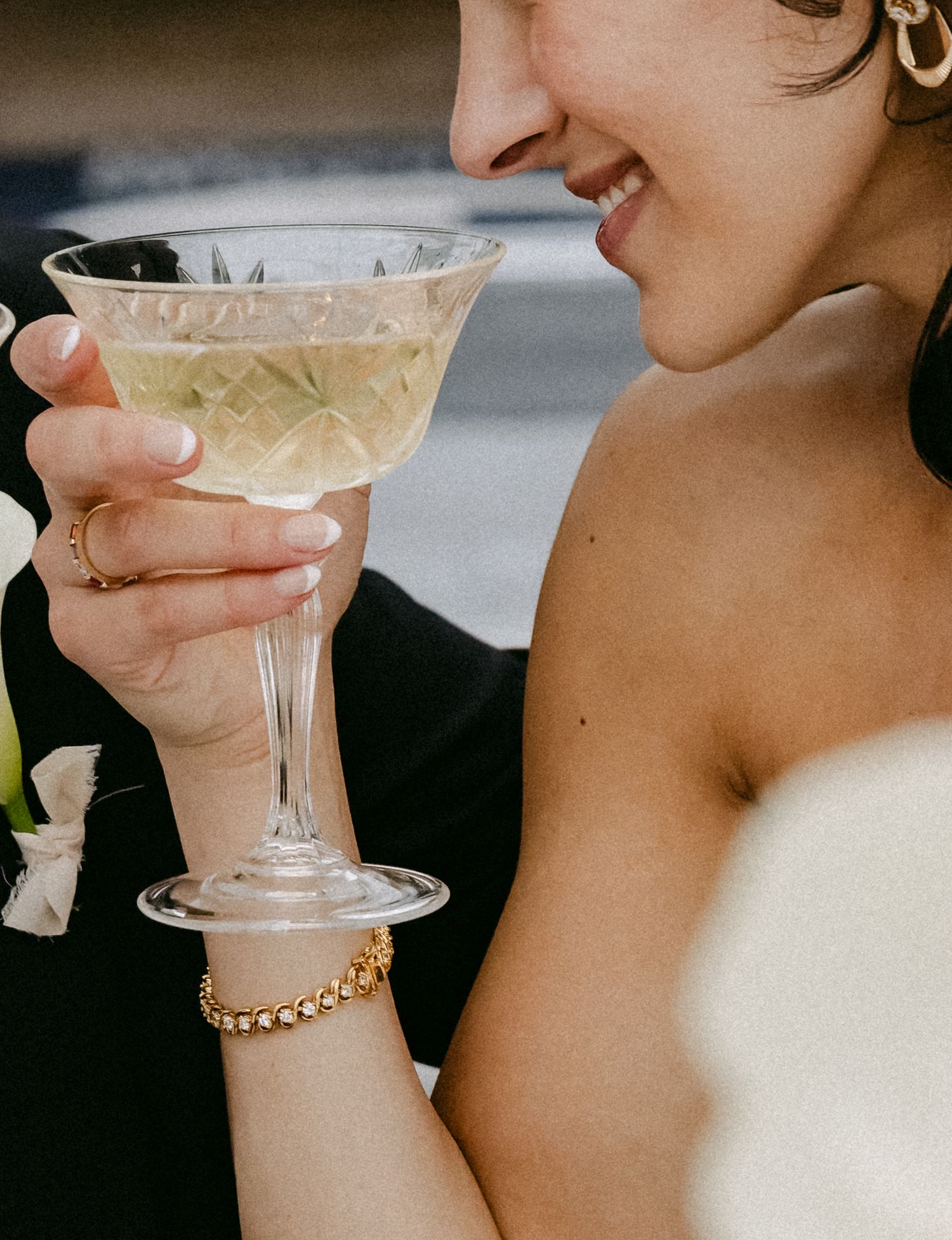 Smiling bride in white dress sipping champagne from crystal coupe glass, wearing gold bracelet and ring at wedding.