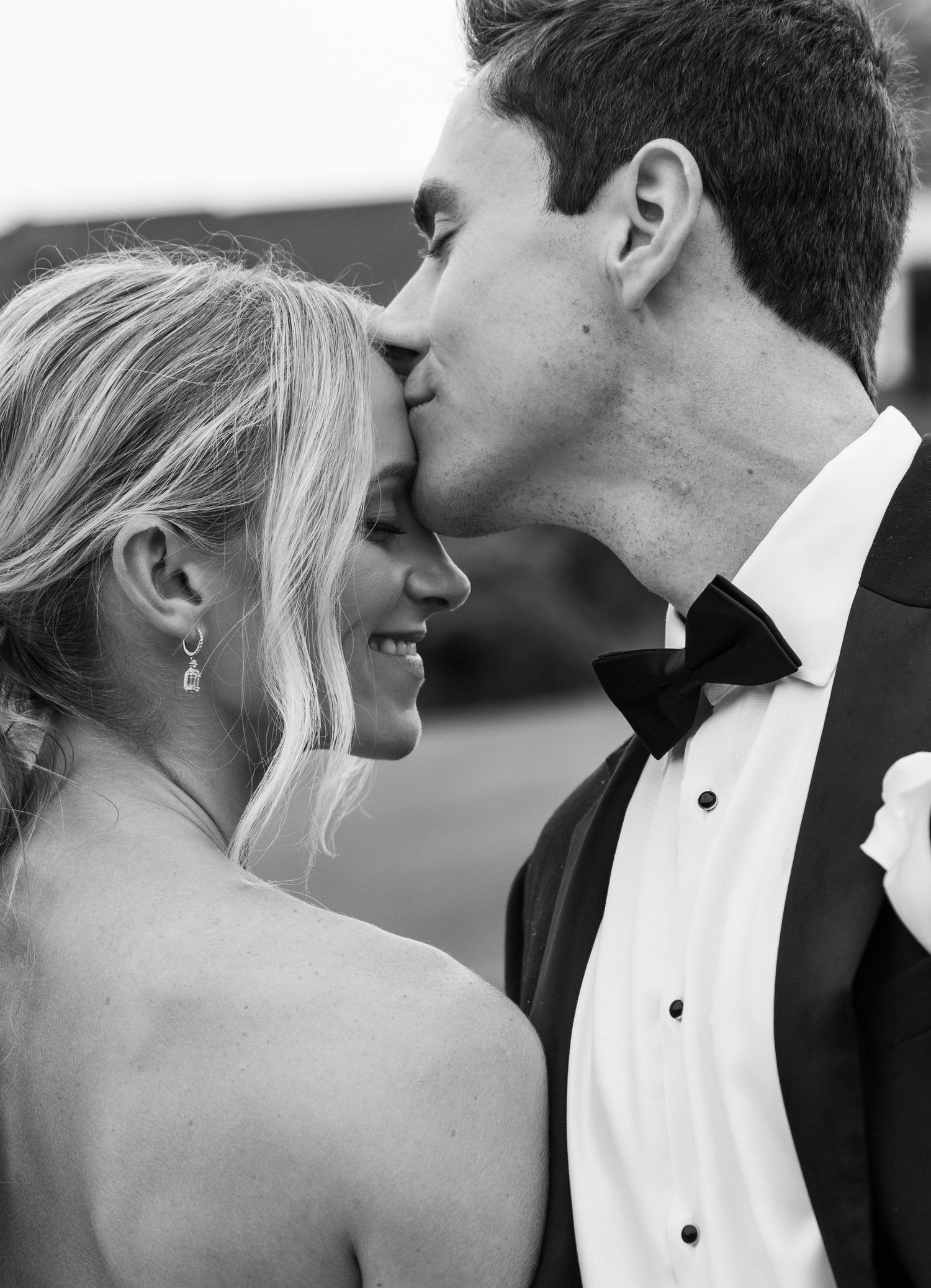 Black and white wedding photo of smiling bride and groom sharing a tender kiss outdoors in formal attire.
