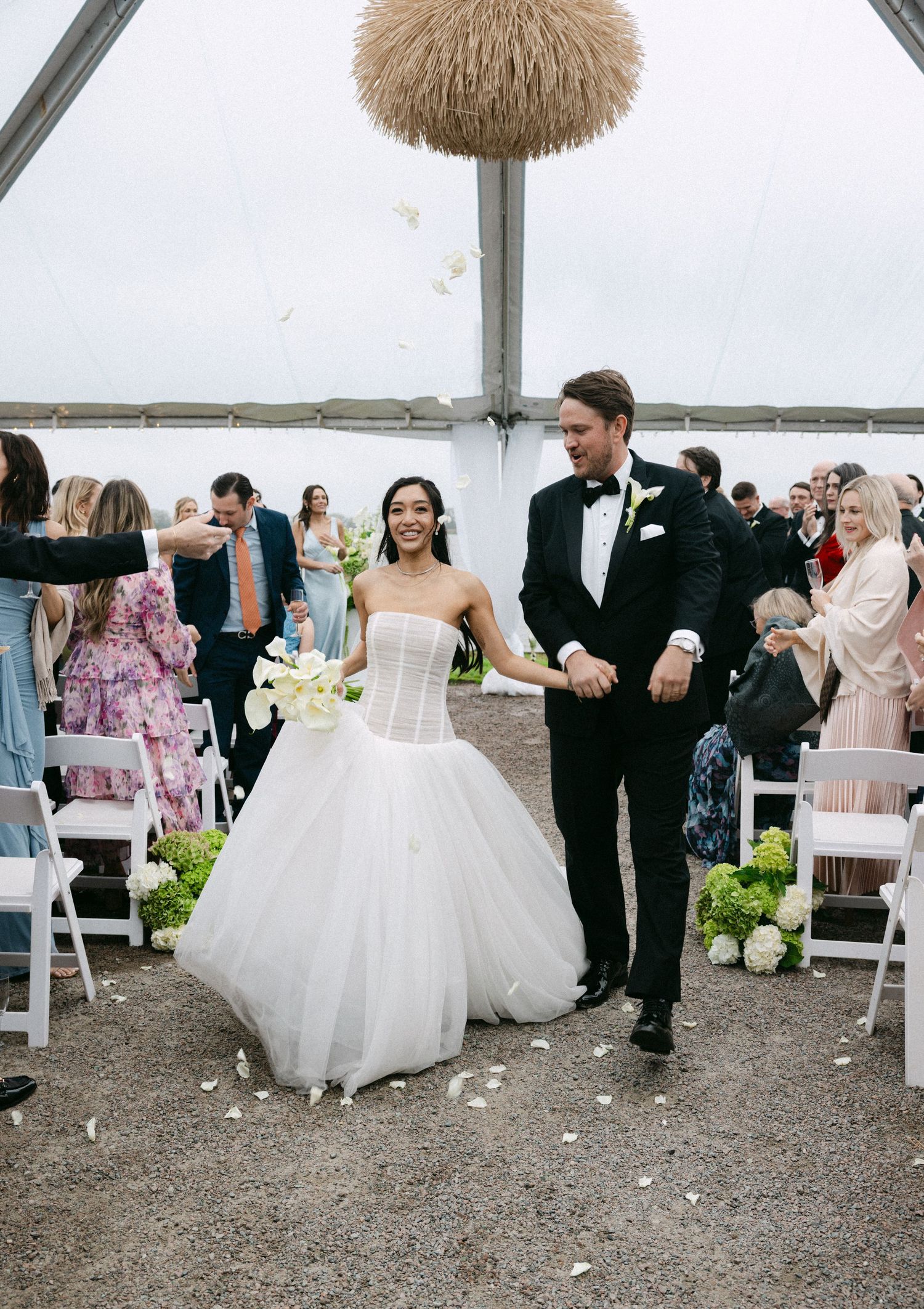 Bride and groom walk down the aisle under a tented outdoor wedding ceremony as guests toss white flower petals.
