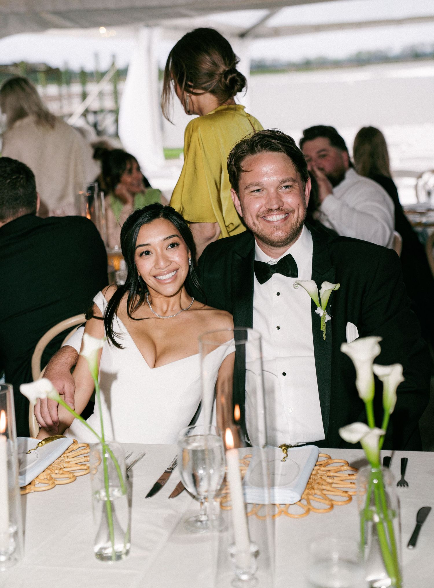 Bride and groom smile at sweetheart table adorned with calla lilies during wedding reception.