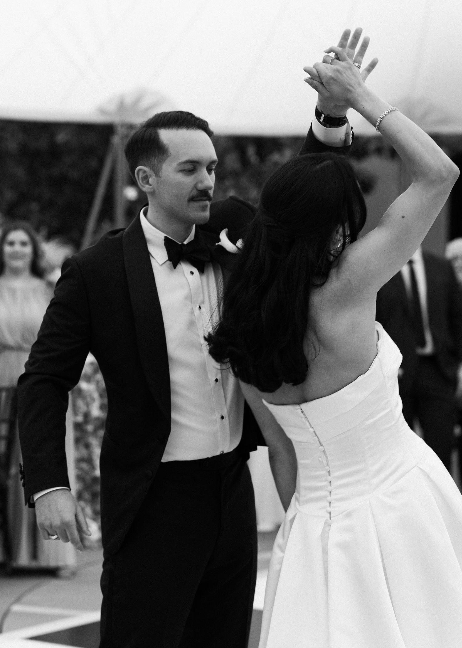 Black and white photo of bride and groom sharing first dance under wedding tent, groom in classic tuxedo.