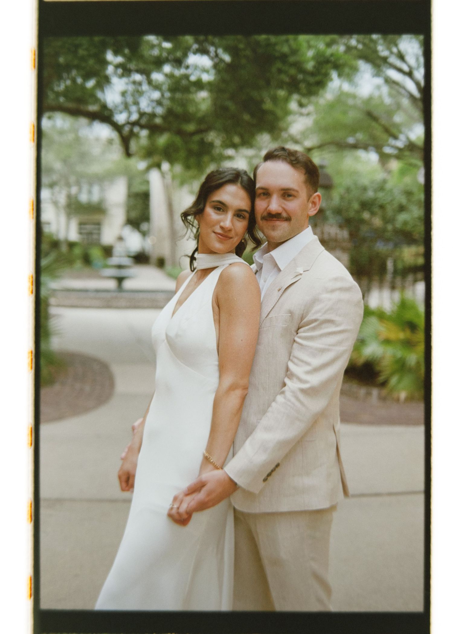 Bride and groom pose together outdoors on film, she in a sleek white gown, he in a beige suit.