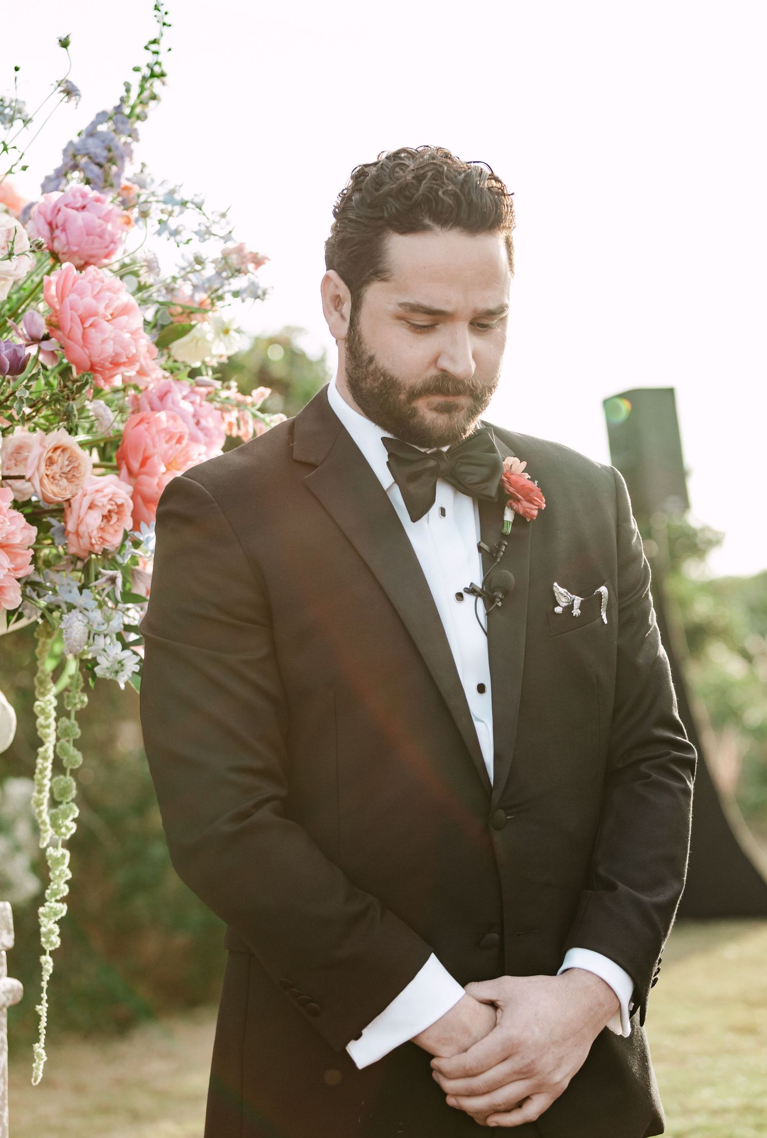 Dapper groom in black tuxedo with bow tie standing near colorful floral wedding ceremony arch.