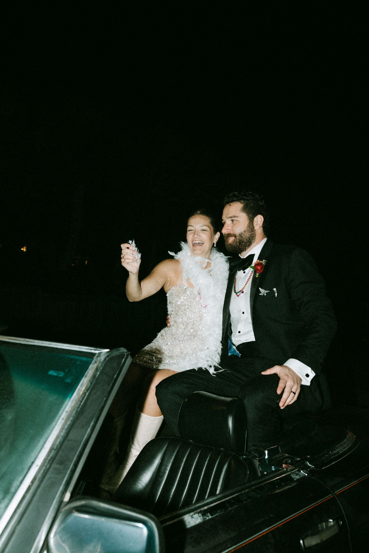 Bride in short feathered dress and groom in tuxedo sit atop vintage convertible car celebrating at night.