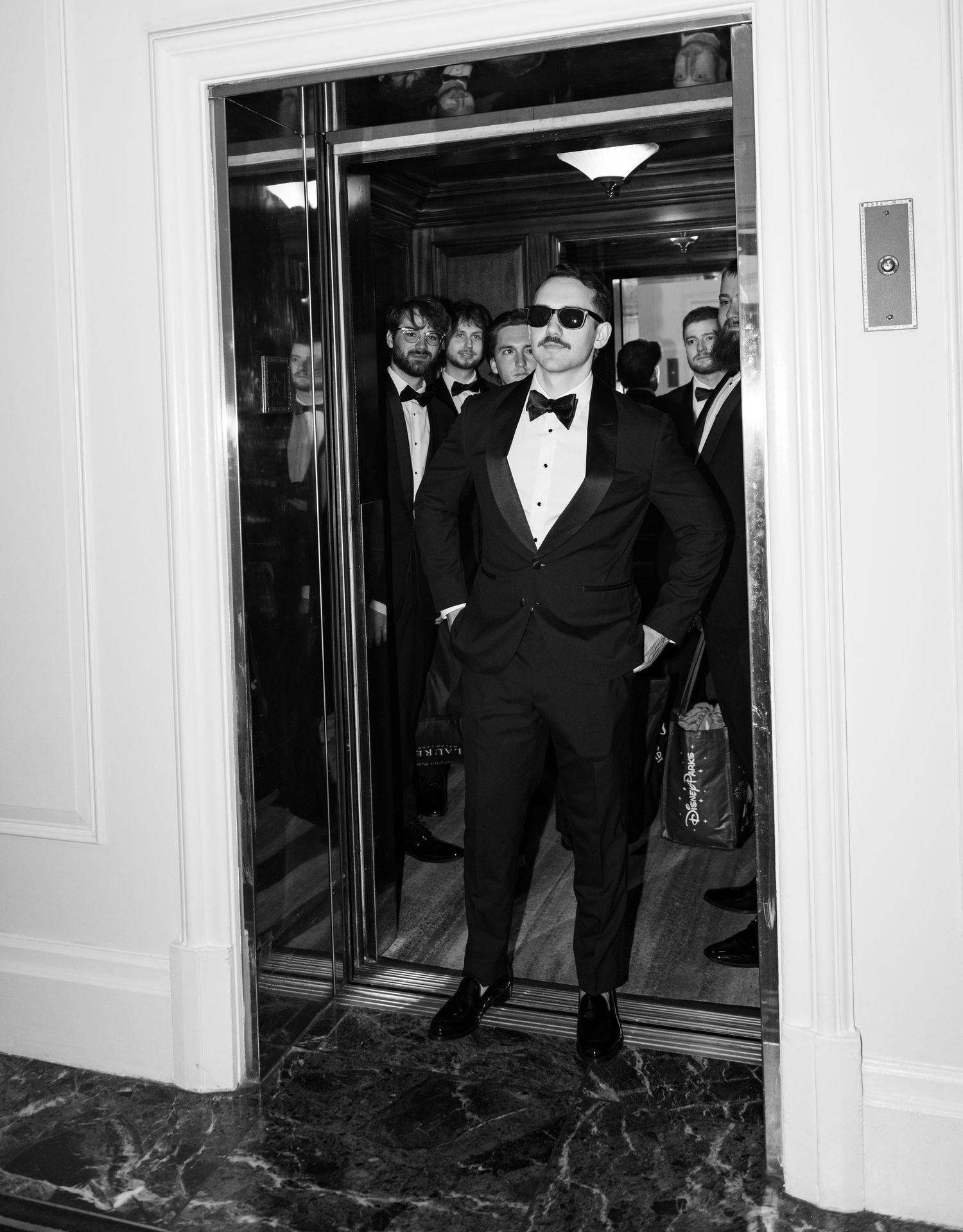Man in tuxedo and sunglasses exits elevator with group in formal wear, black and white photo.