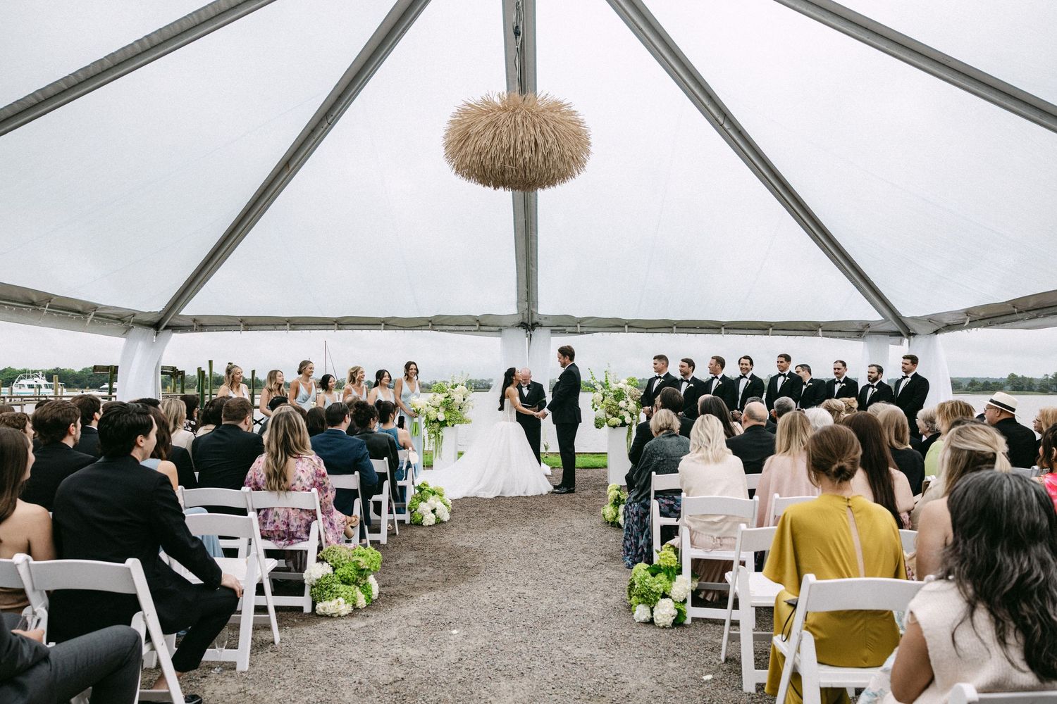 Elegant outdoor wedding ceremony under a clear tent with white chairs, floral arrangements, and a chandelier overlooking a waterfront.