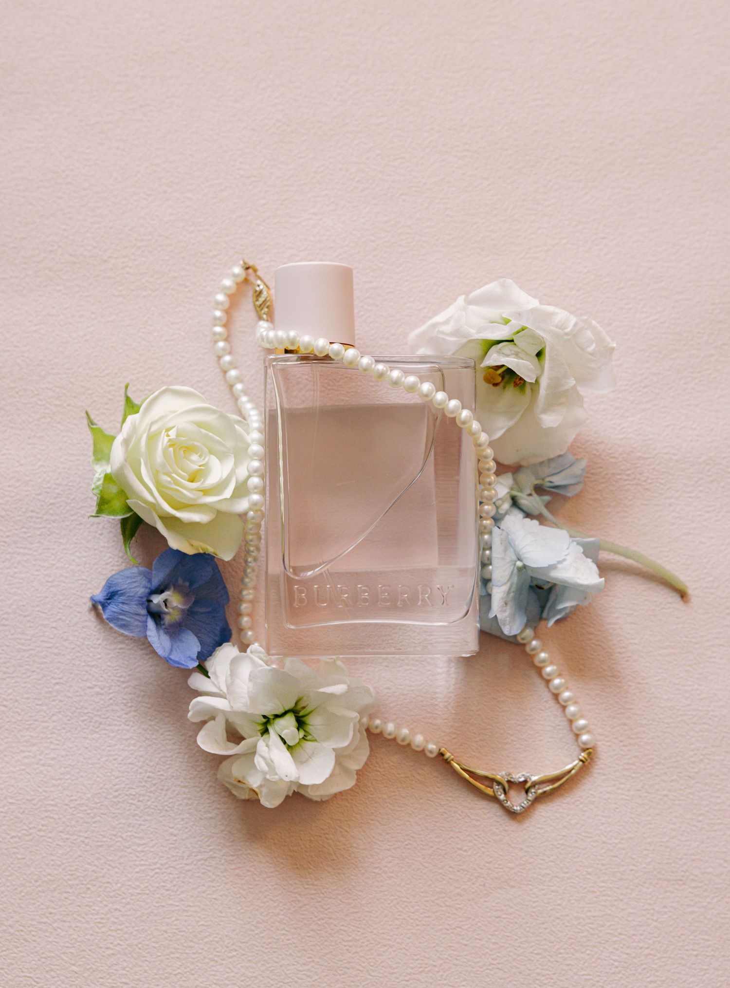Burberry perfume bottle surrounded by white and blue flowers with a delicate pearl necklace on a soft pink background.