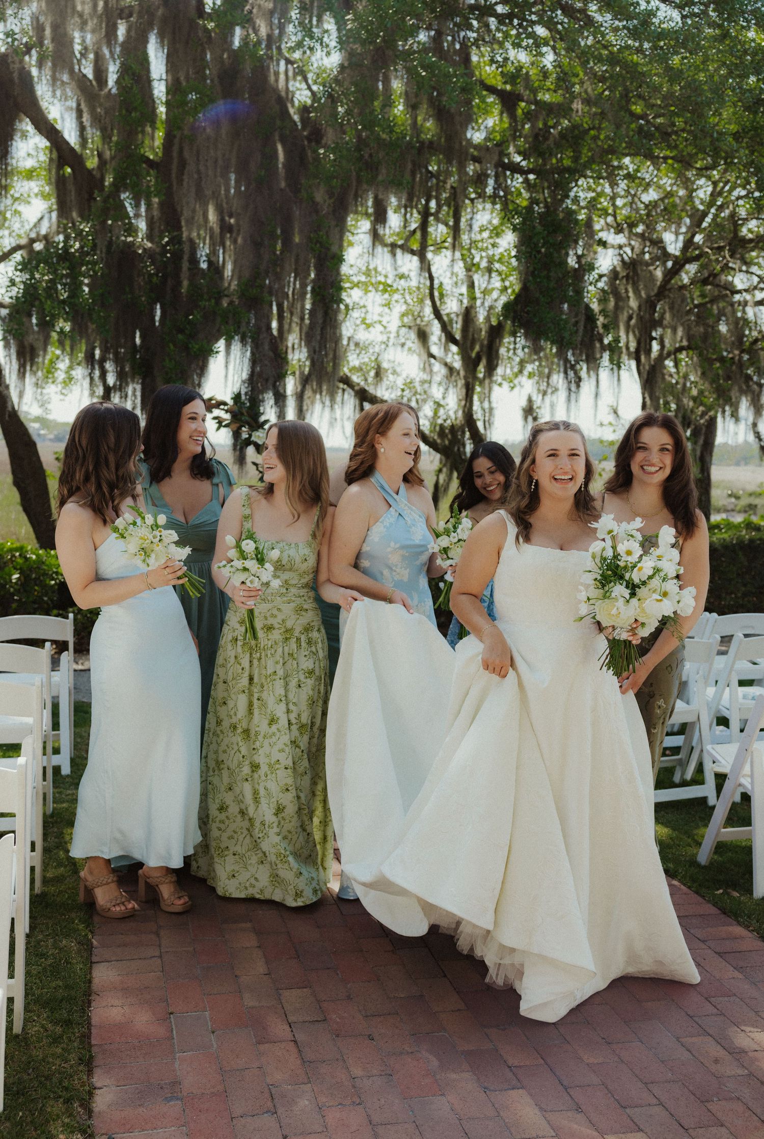 Bridesmaids in light-colored dresses pose together under moss-draped trees at an outdoor venue.