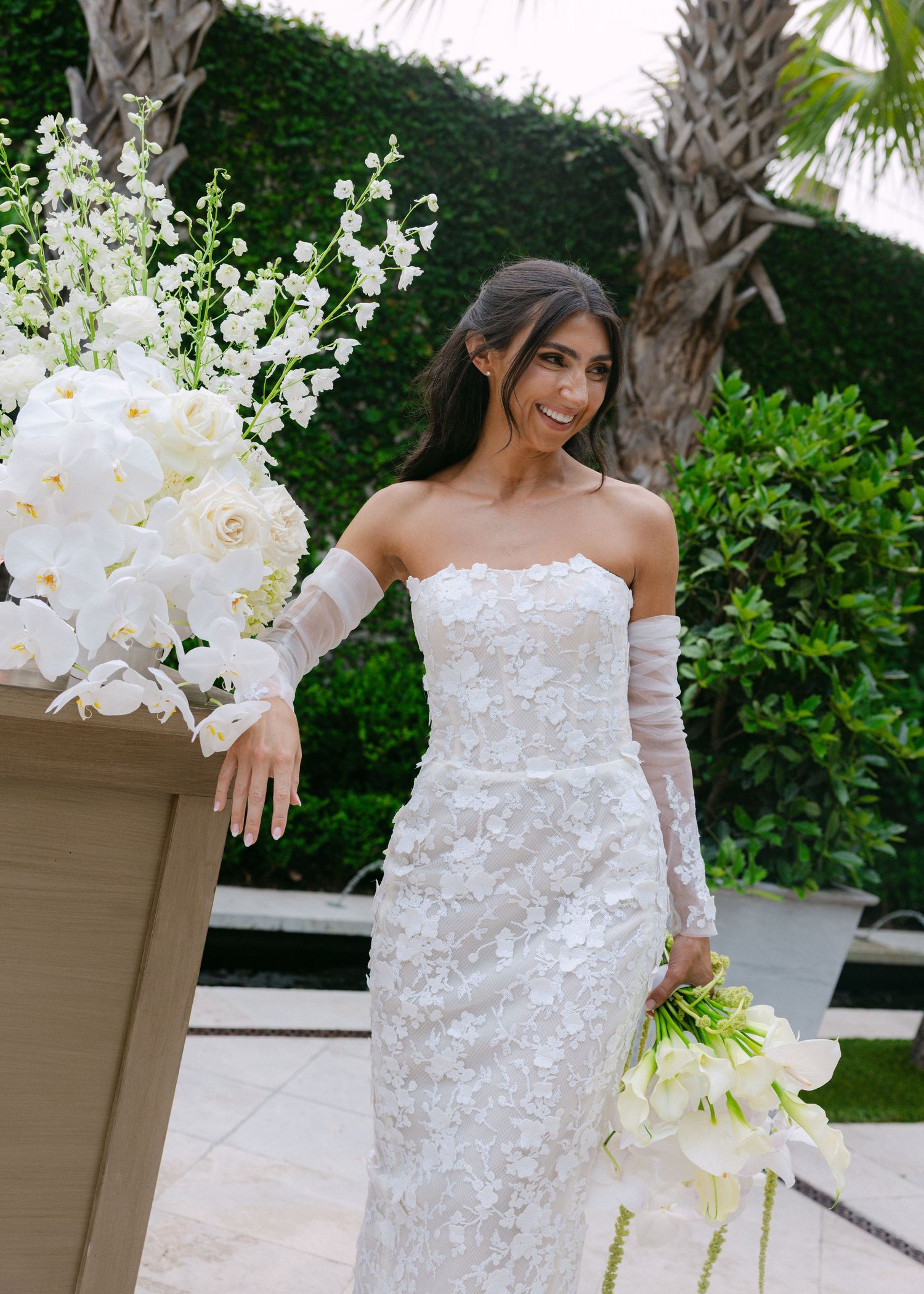 Bride in strapless lace gown shows off her wedding dress while holding white orchids in a garden setting.