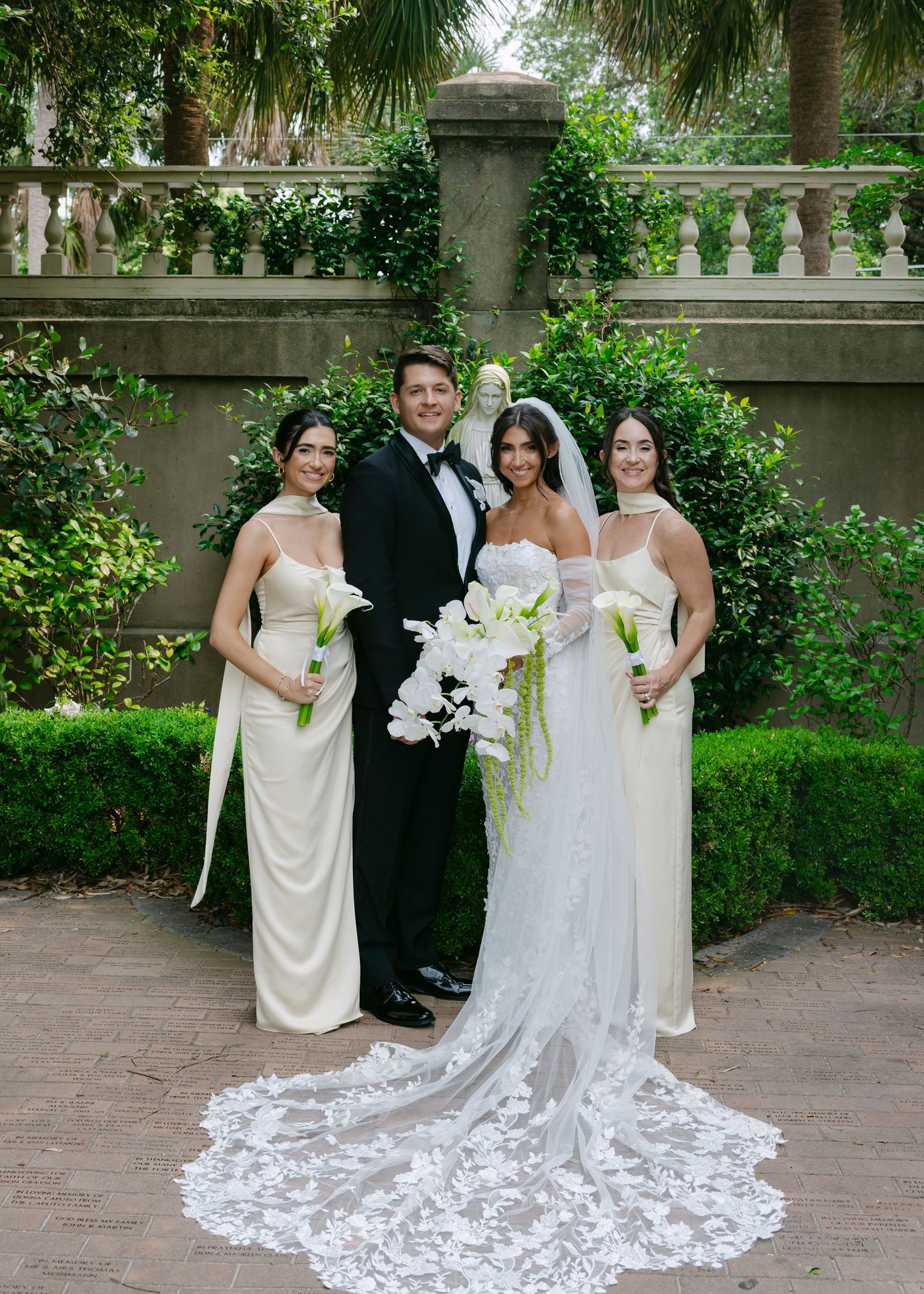 A wedding party poses in a garden setting with a stunning lace train dress in focus.