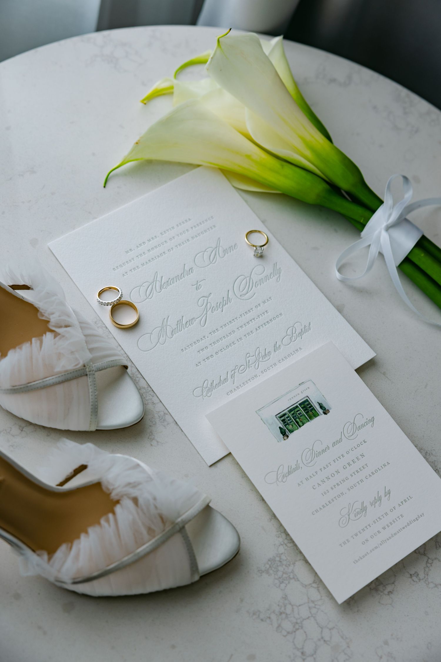 Wedding invitation with white calla lilies and wedding rings displayed on marble surface.