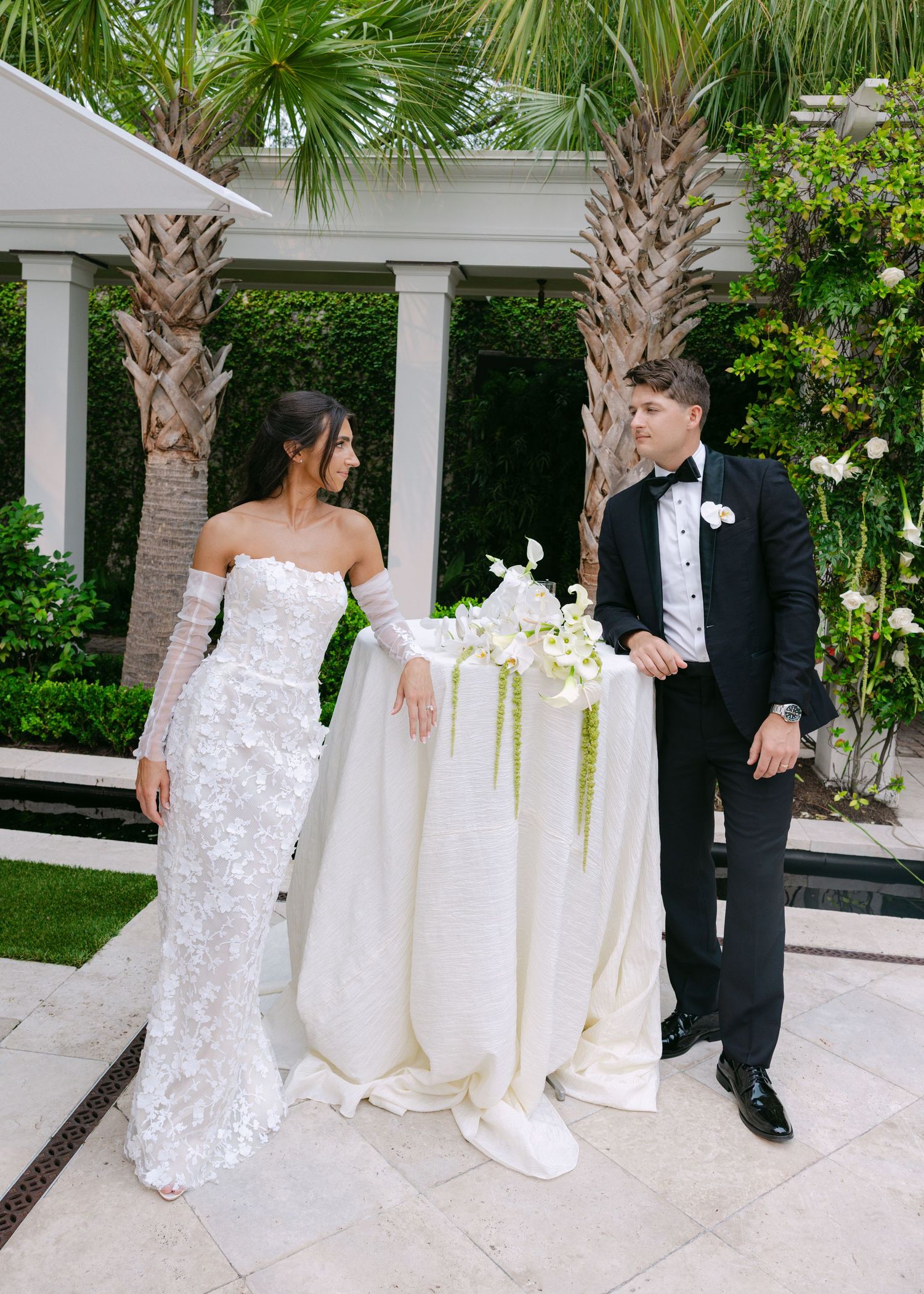Wedding portraits by white columns and palm trees featuring bride in lace gown and groom in tuxedo.