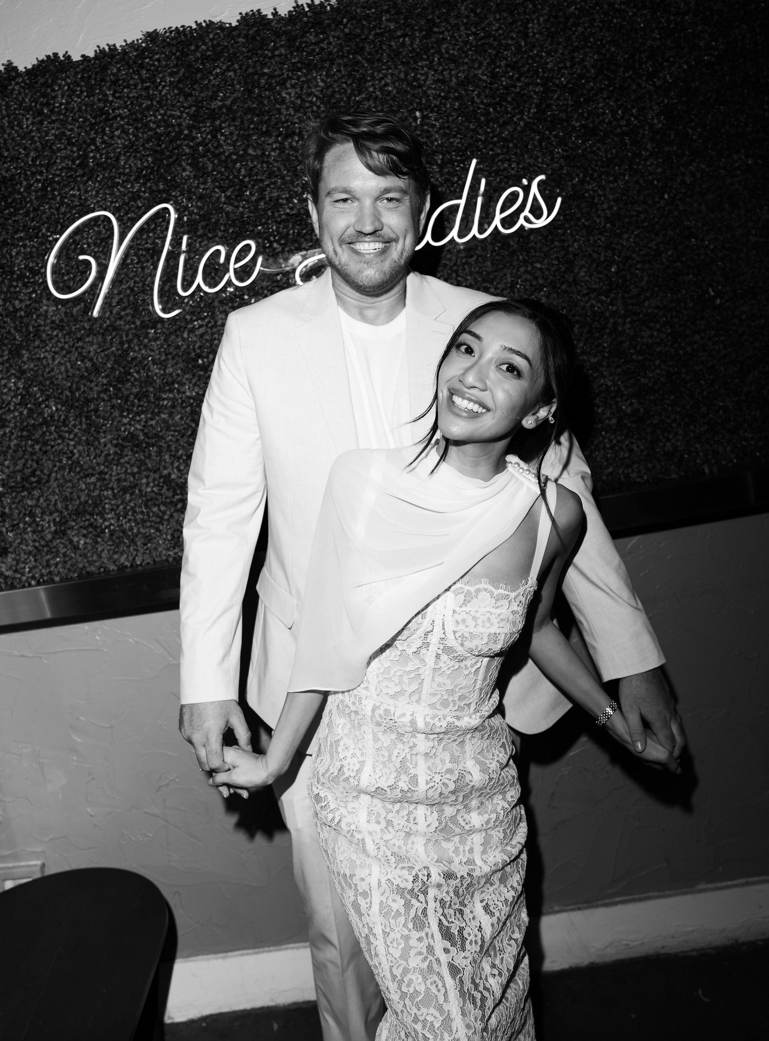 A smiling couple poses together in front of a neon sign at an elegant black and white event, dressed in formal white attire.