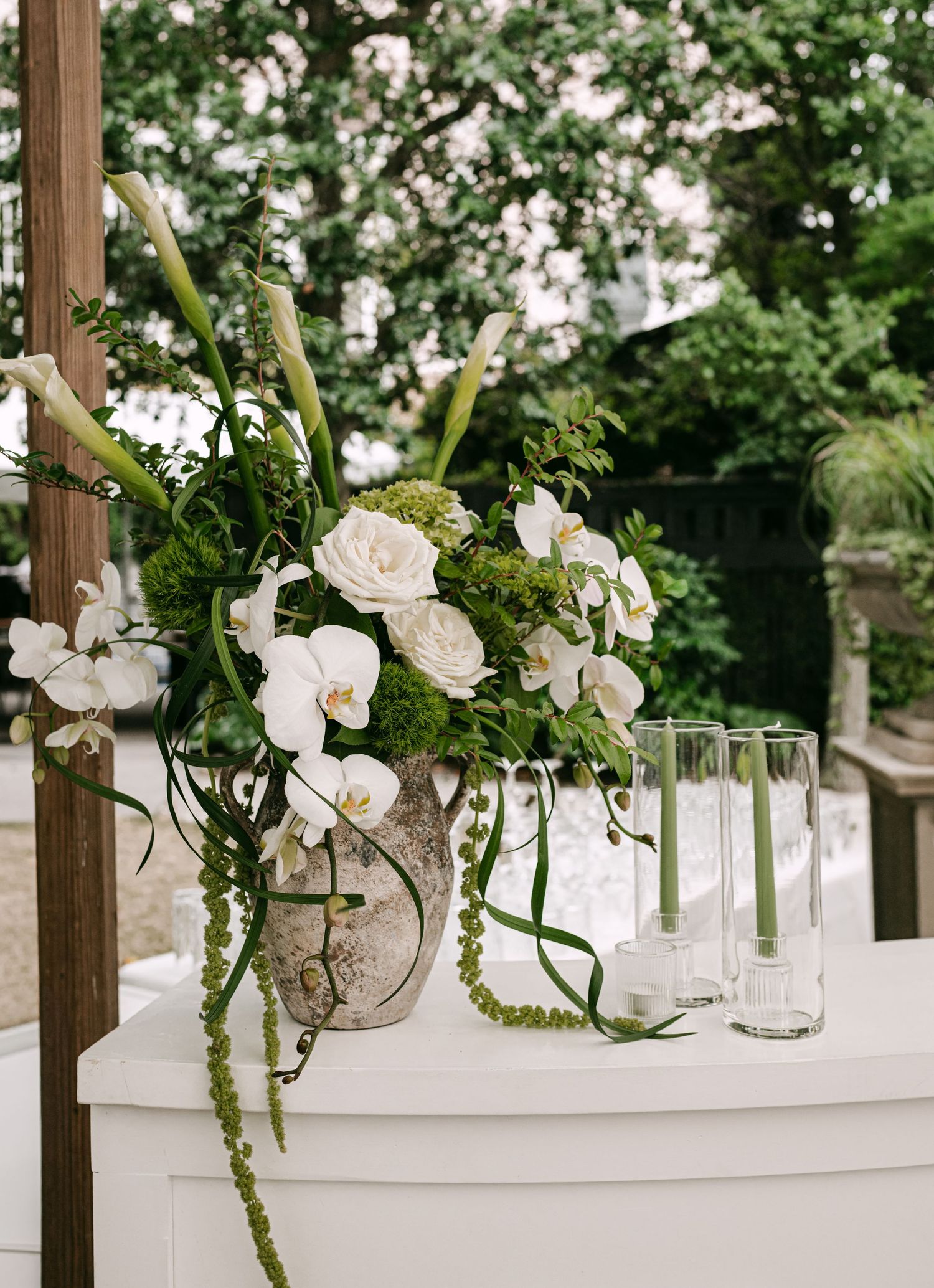 Elegant wedding floral arrangement with white orchids, roses, and greenery in a stone vase beside glass candle holders.