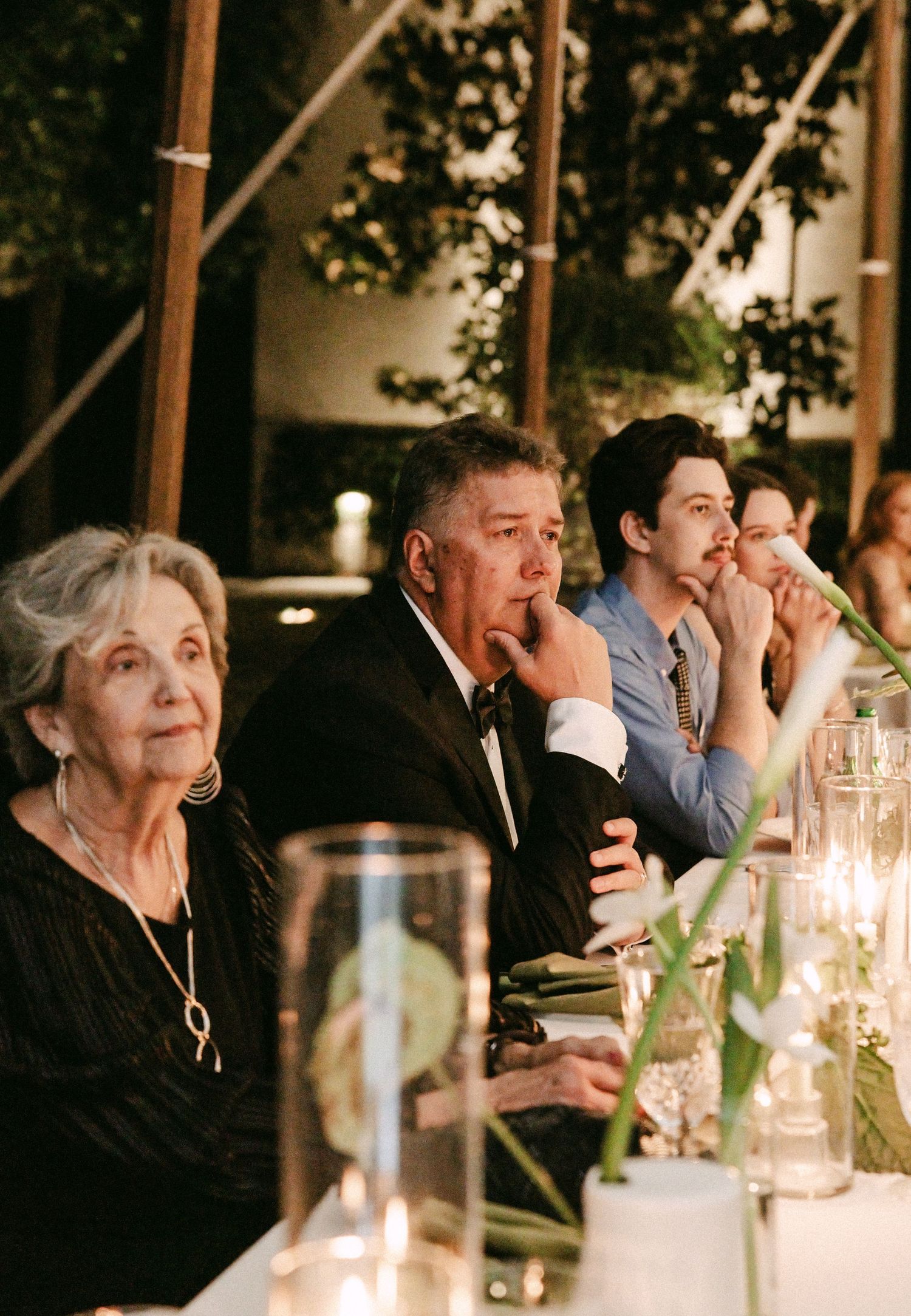 Wedding guests attentively listening to speeches at a candlelit outdoor reception under a tent at night.