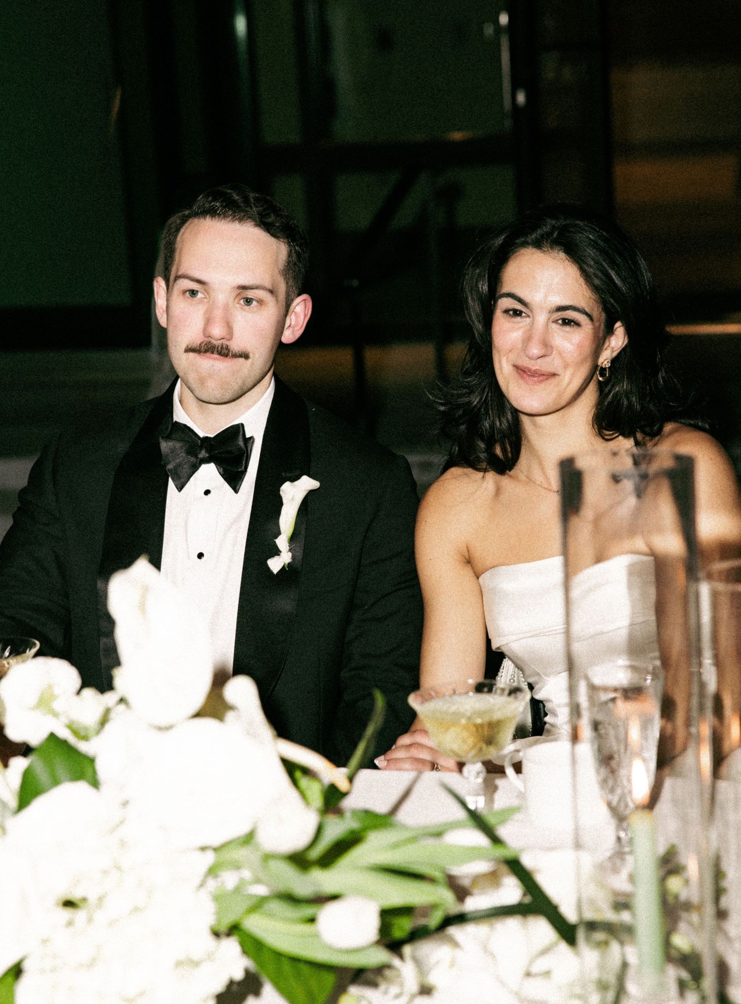 Bride and groom in black tuxedo seated at wedding reception table with white floral arrangements in foreground.
