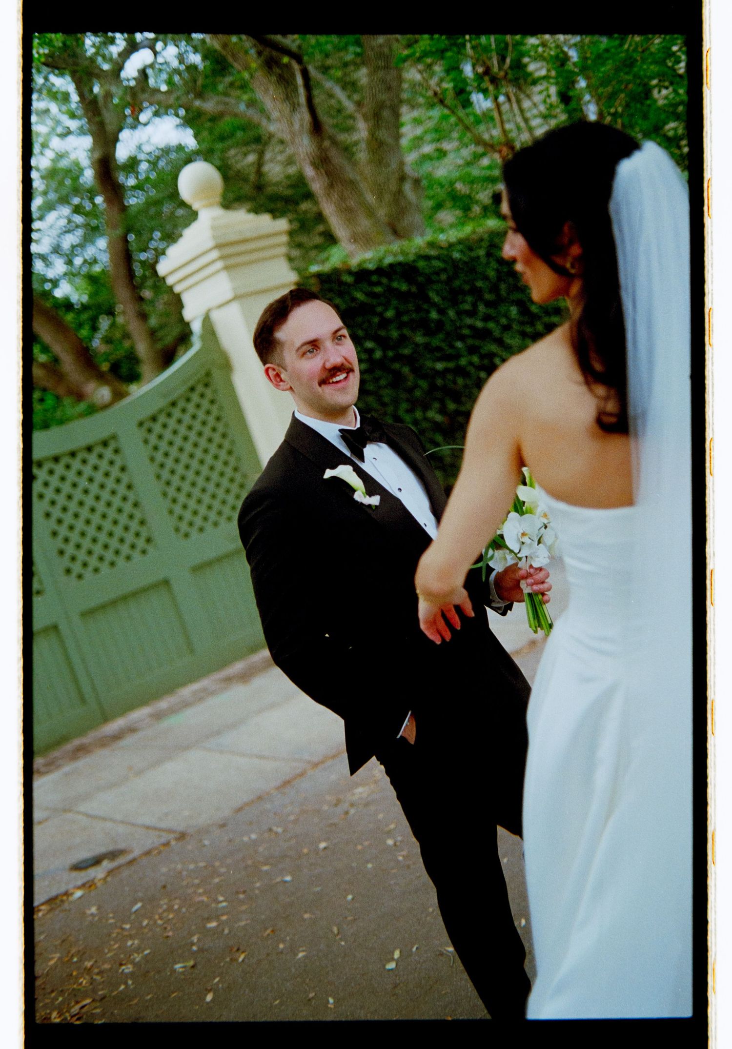 Smiling groom in black tuxedo looks back at bride in white gown and veil during outdoor wedding portraits.