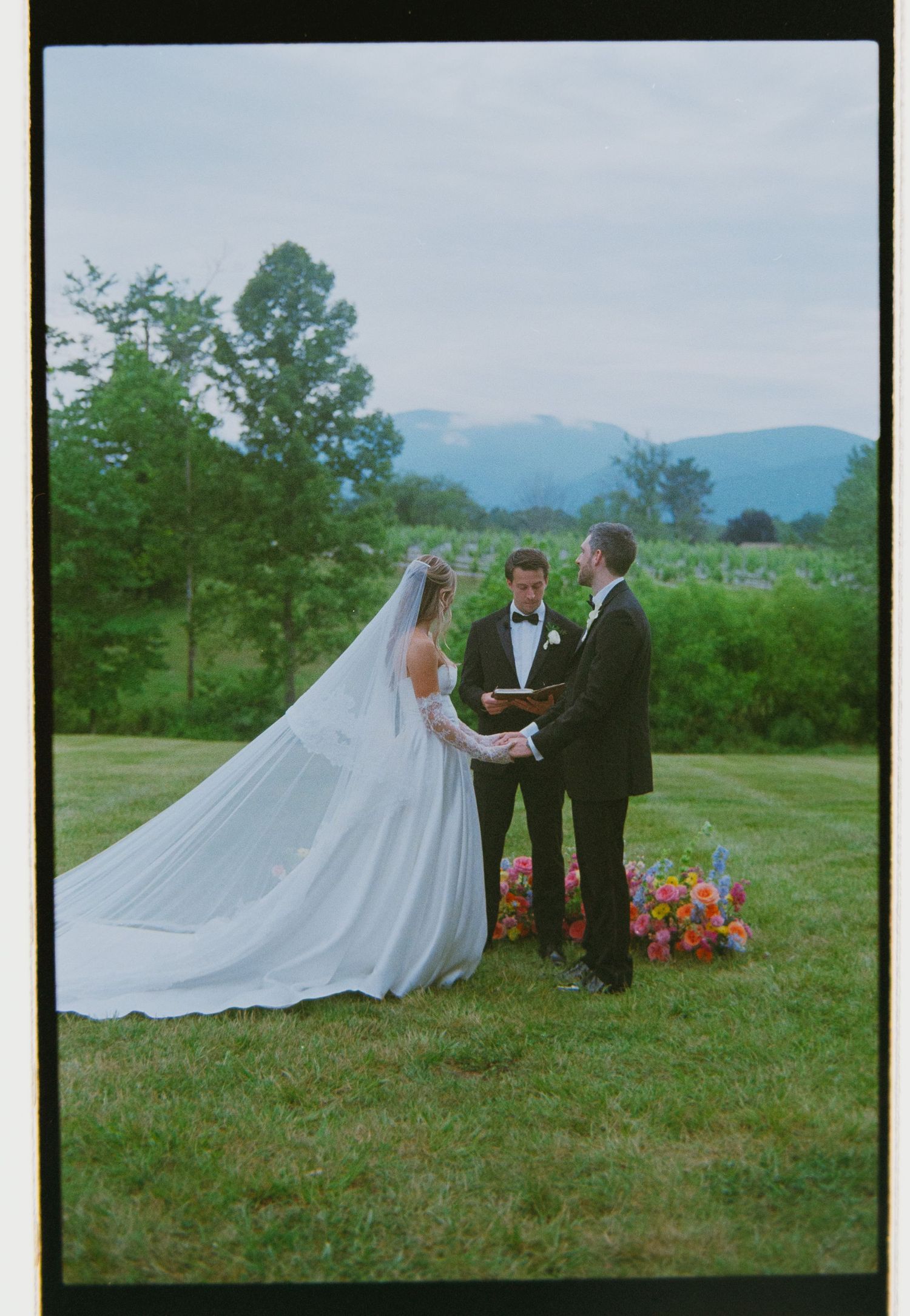 Bride and groom exchanging vows outdoors with mountain views, officiant holding book, colorful flowers, long cathedral veil.