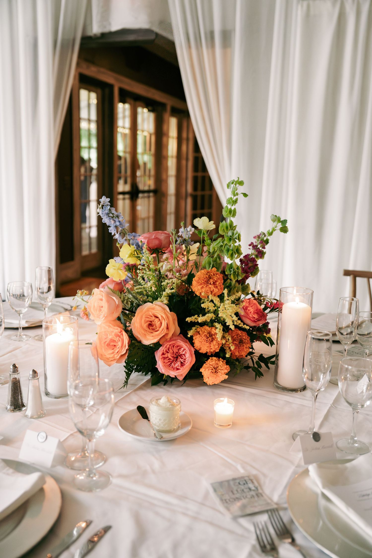 Colorful floral centerpiece with orange and pink roses surrounded by candles on white reception table.