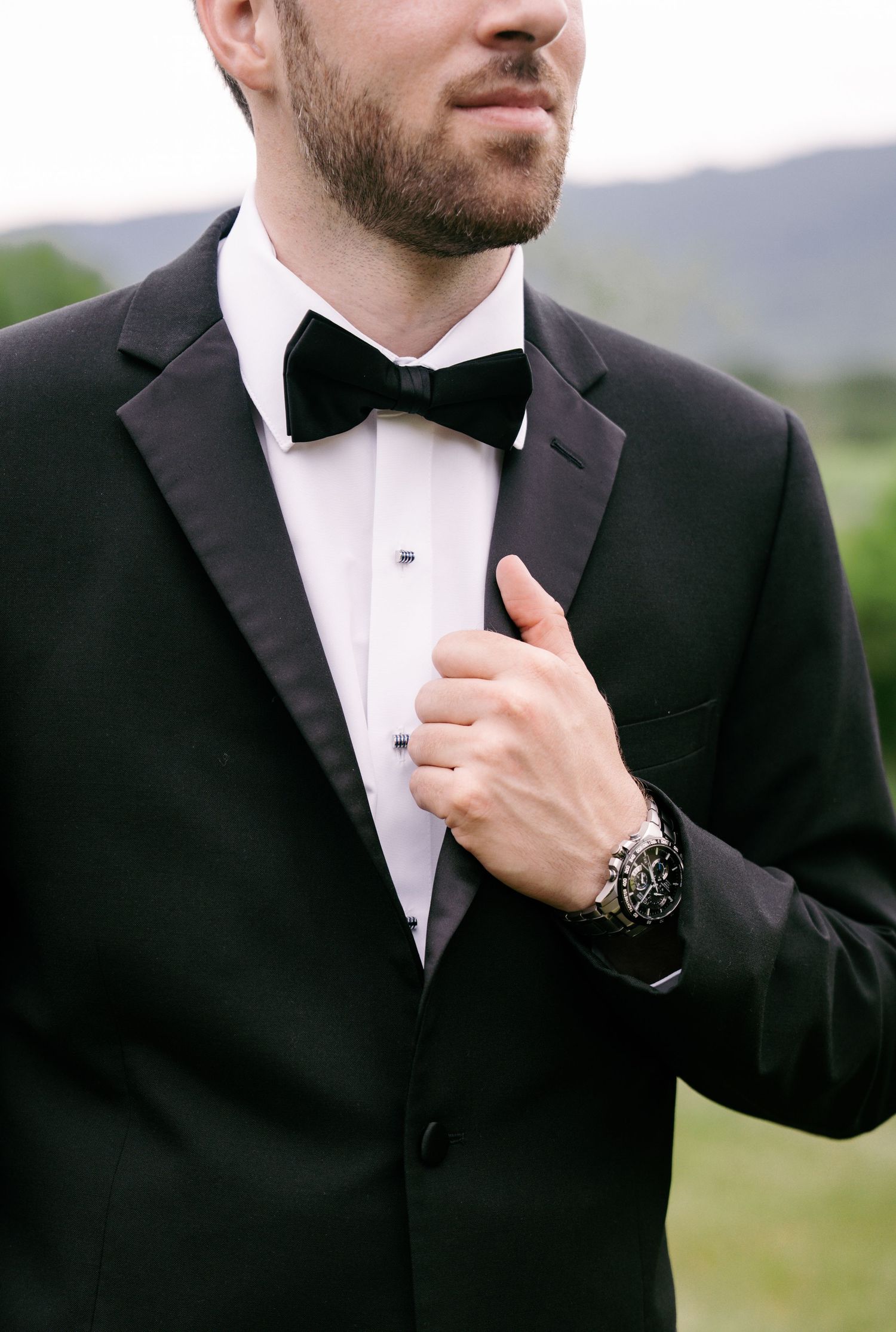 Groom in black tuxedo with velvet bow tie and dress watch adjusting his jacket lapel outdoors.