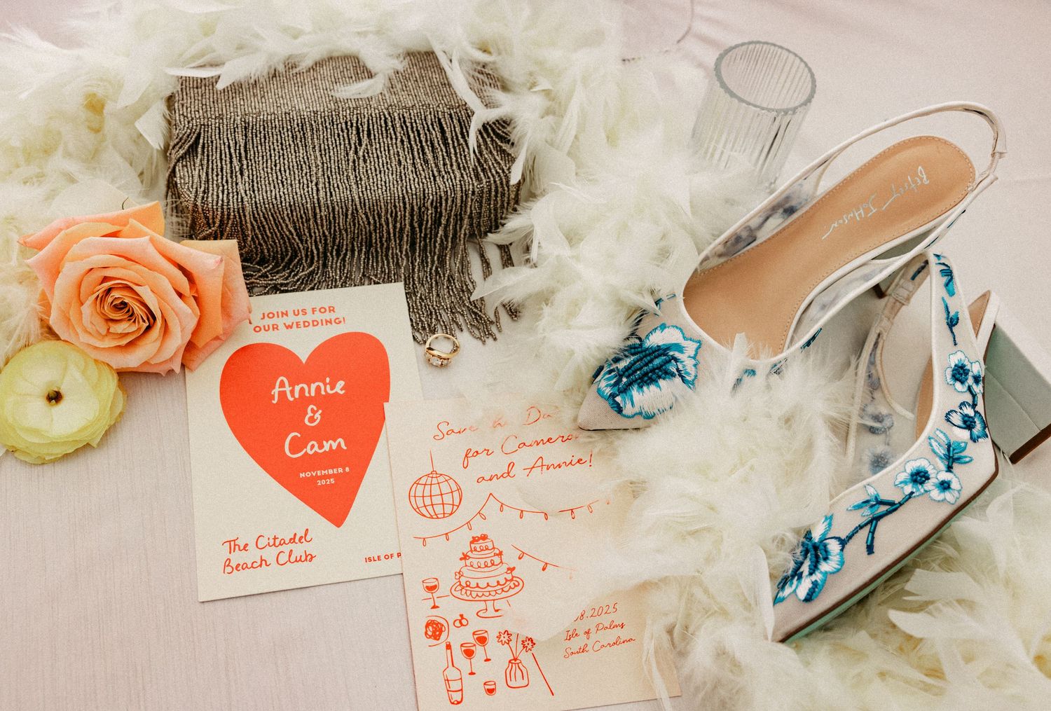 Elegant wedding flat lay with embroidered heels, coral roses, fringe clutch, and Annie & Cam invitation on white feathers.
