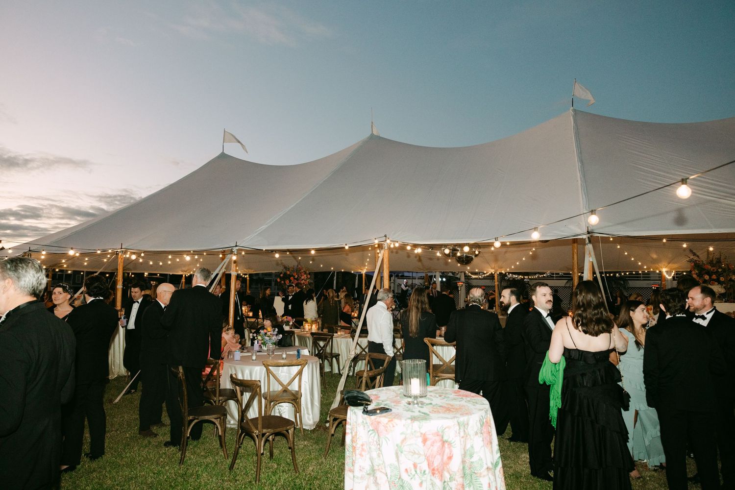Guests mingle under a large white sailcloth tent with string lights at evening wedding reception.