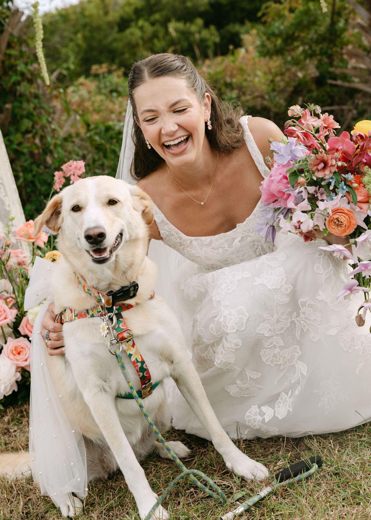 Laughing bride in white lace gown poses with yellow Labrador dog amid colorful floral arrangements outdoors.