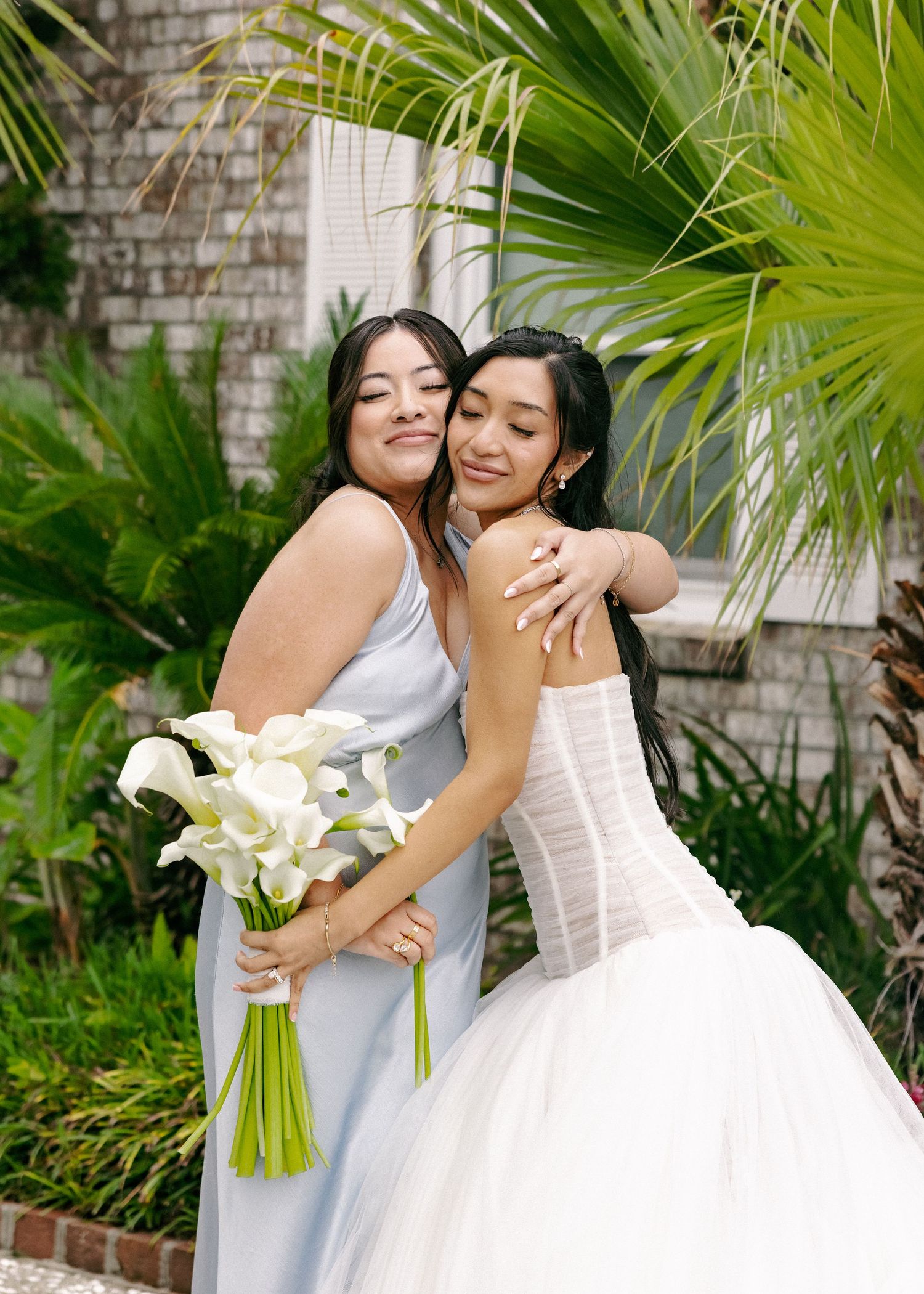 Bride in strapless corset gown embraces bridesmaid in blue dress holding white calla lily bouquet outdoors.