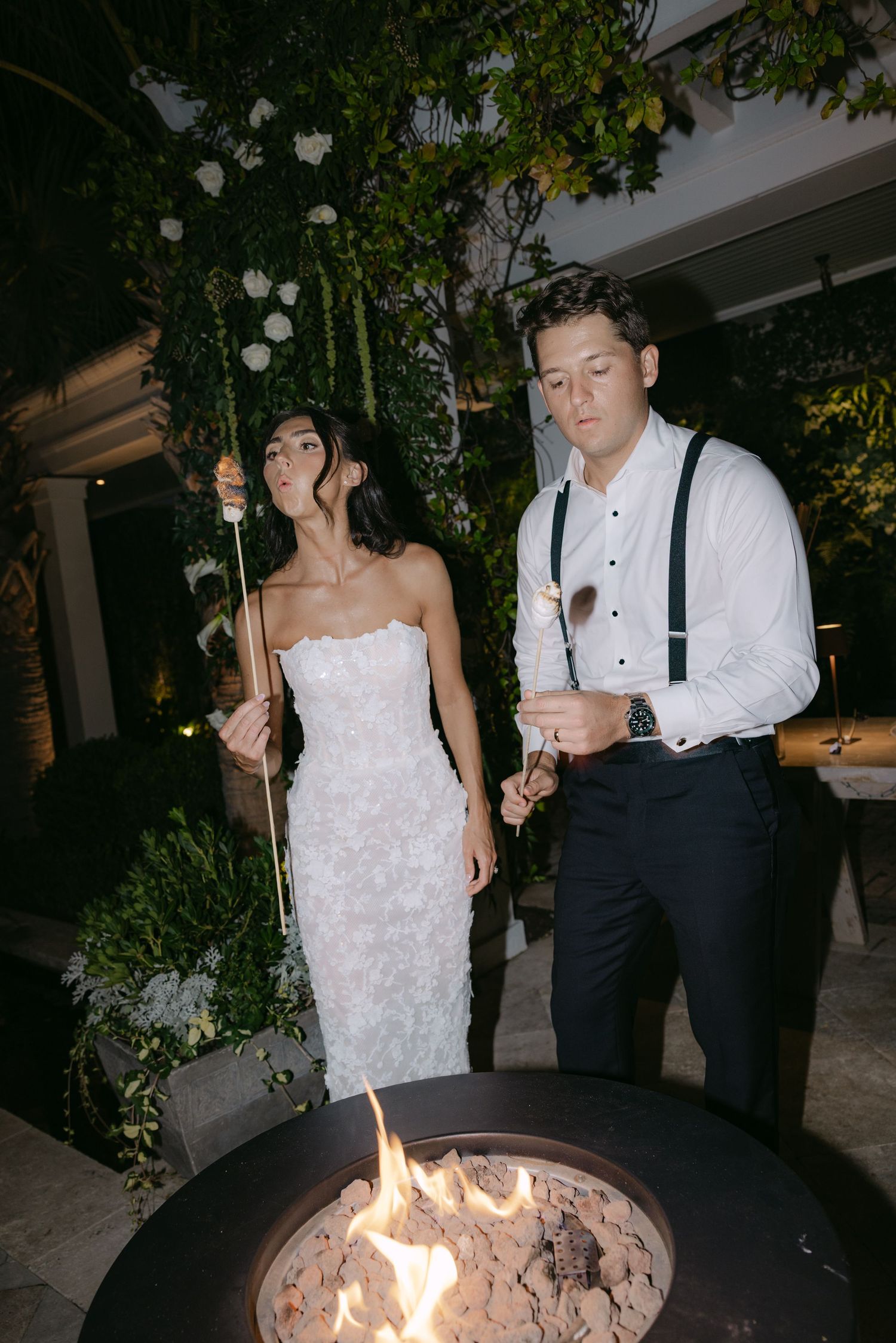 Couple making s'mores by fire pit at elegant outdoor wedding reception.