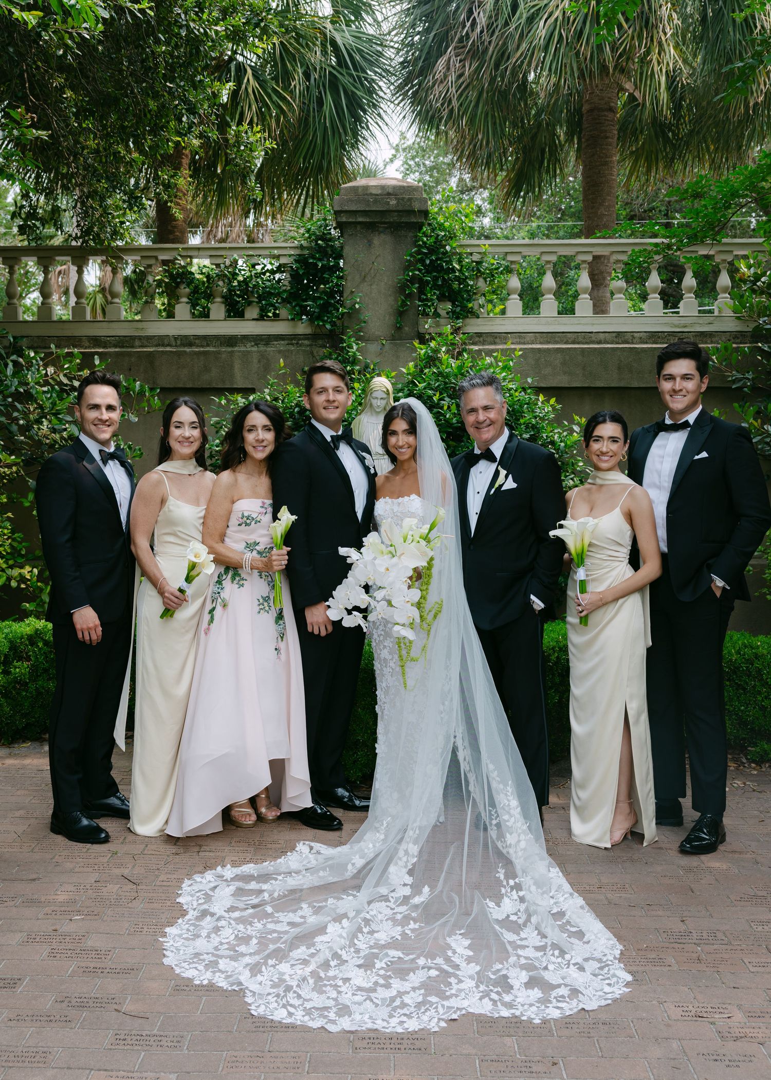 Formal wedding party in cream and black attire poses together in tropical garden setting.