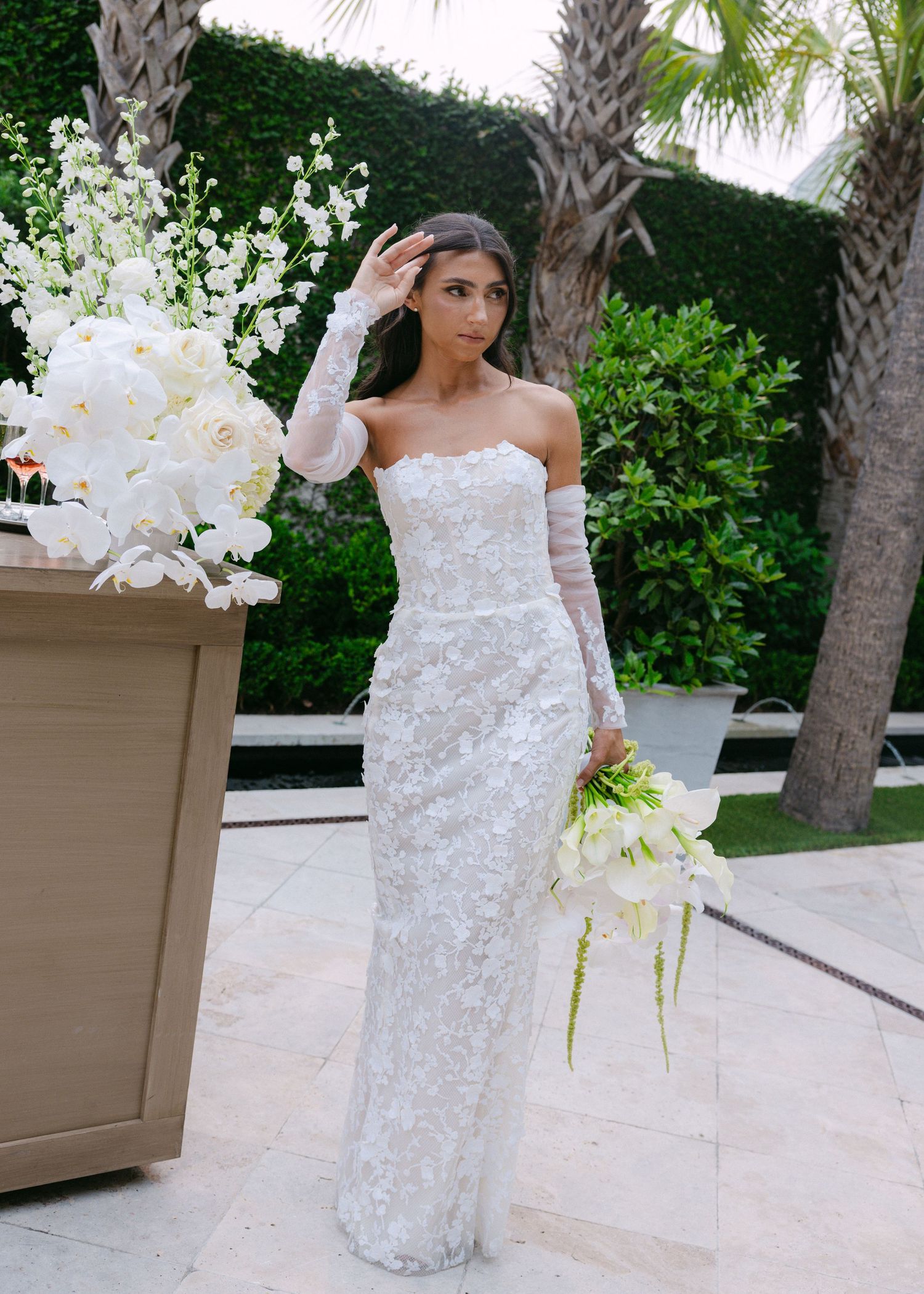 Bride in stunning off-shoulder lace gown holds elegant white orchid bouquet against tropical garden backdrop.