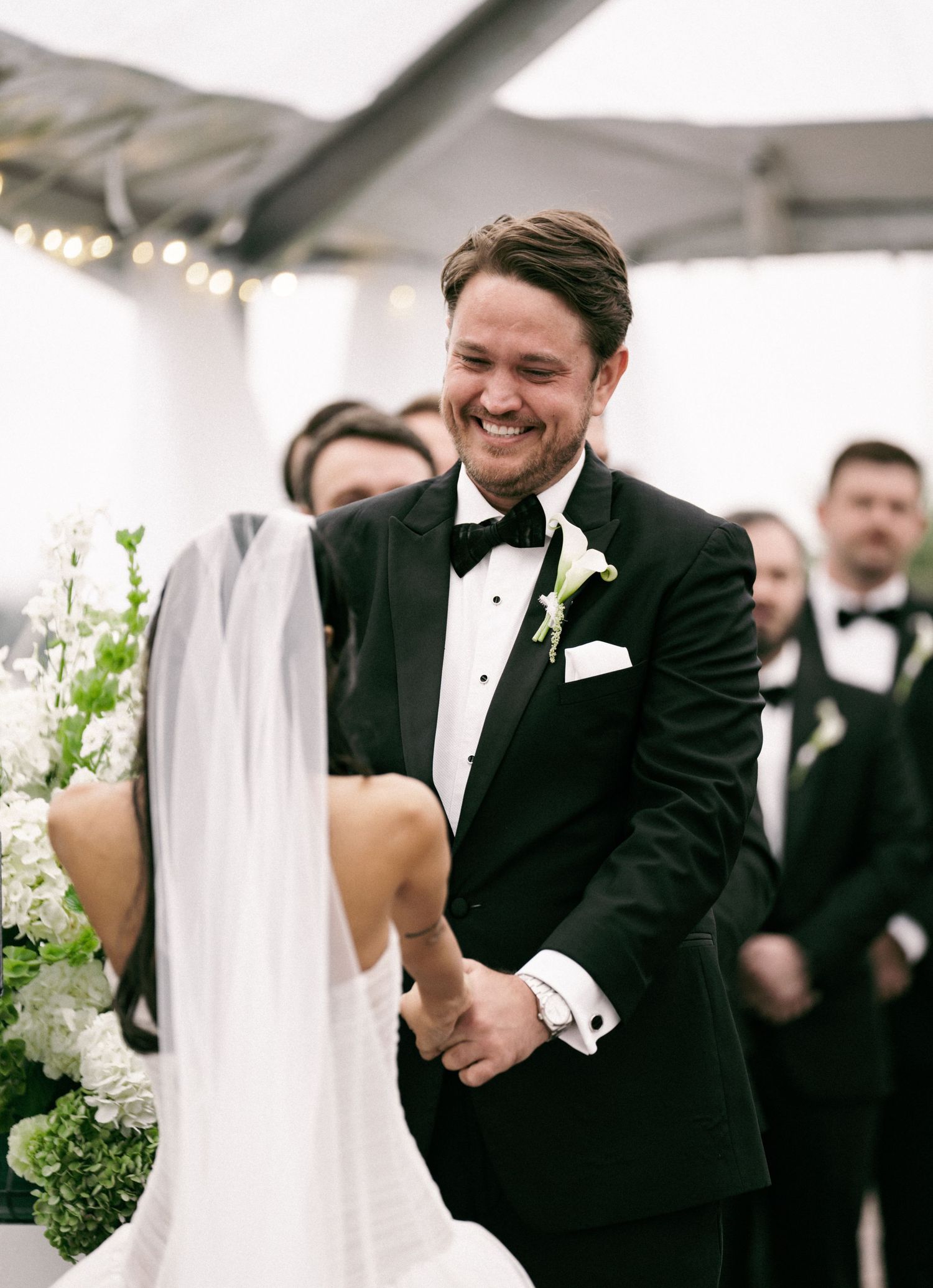 Groom in black tuxedo smiling joyfully at bride during outdoor wedding ceremony with white floral arrangements.