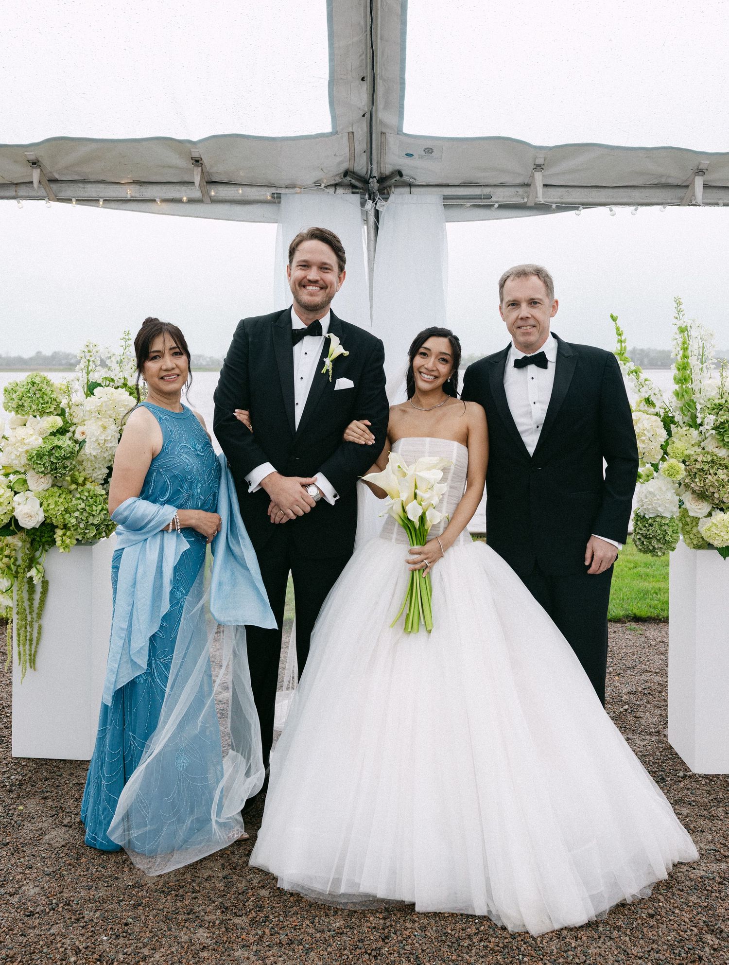 Wedding party posing under tent with bride in ball gown holding calla lilies, groom in tuxedo, and two guests in formal attire.