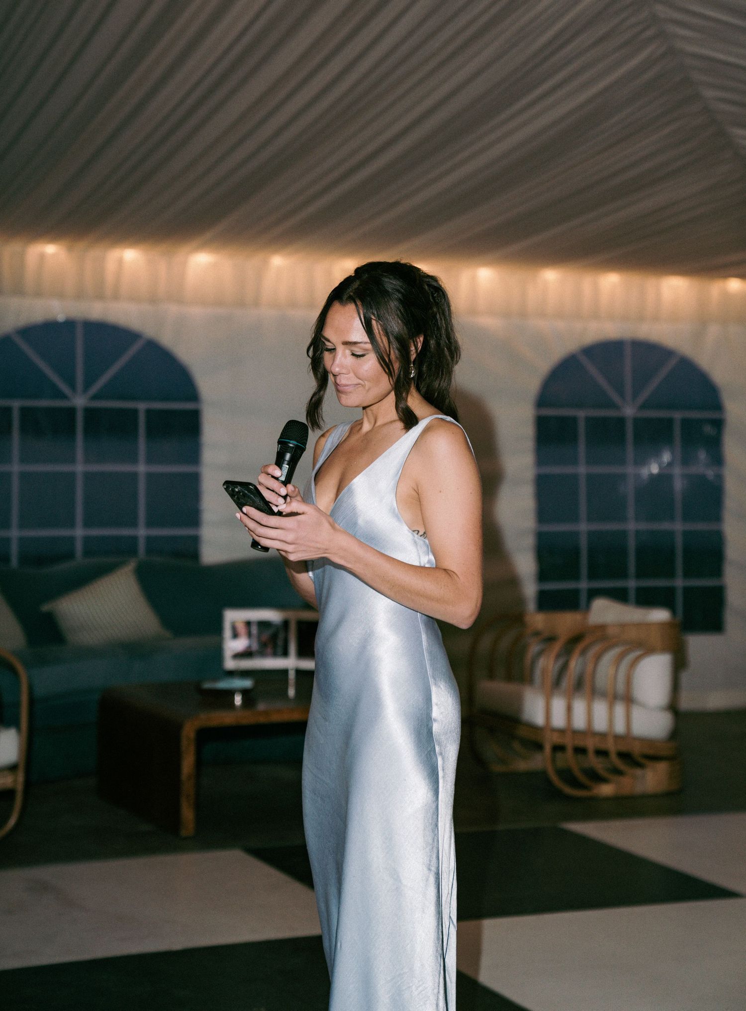 Elegant woman in silver slip dress giving a speech at a wedding reception while reading from her phone.
