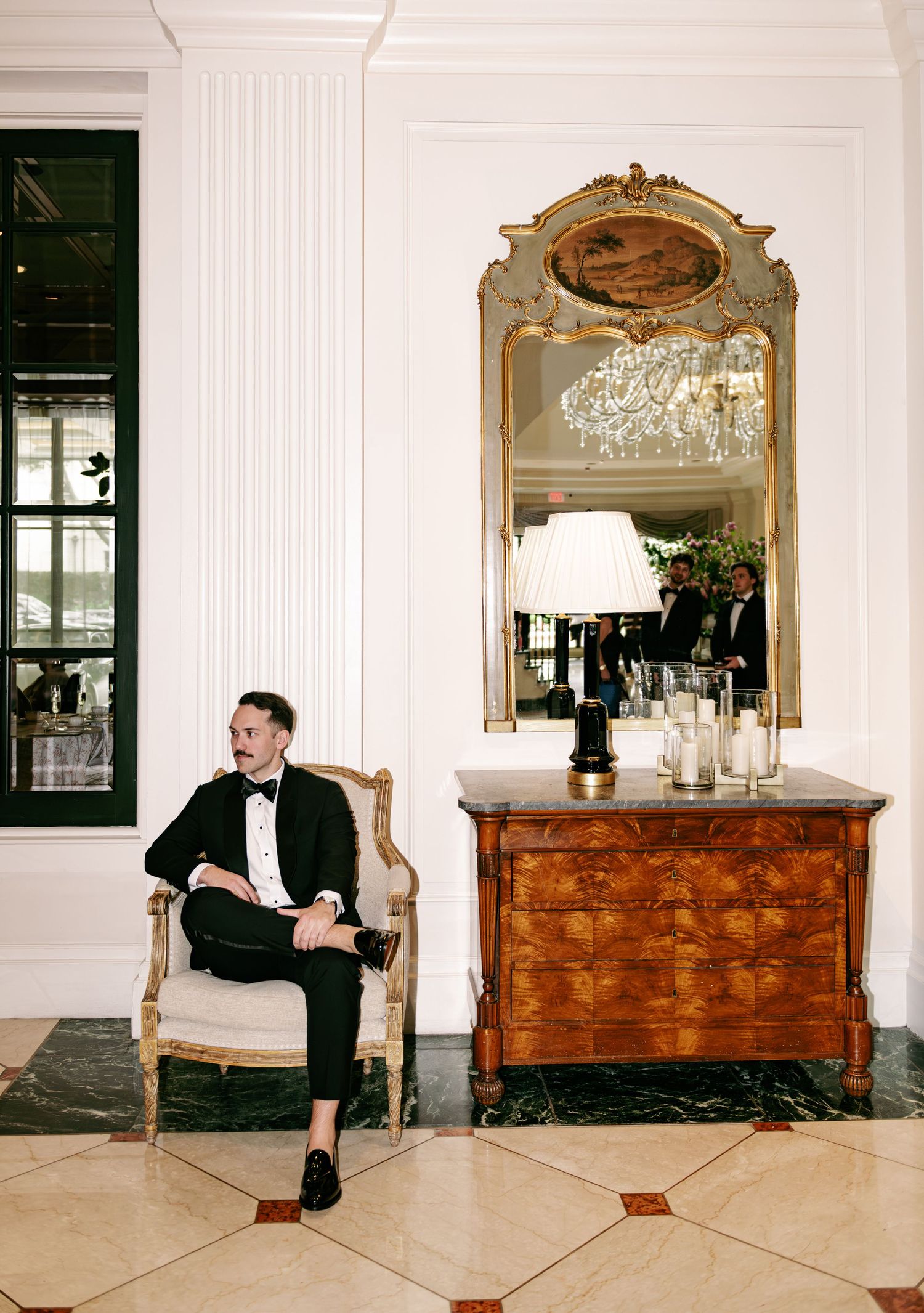 Man in black tuxedo sitting in ornate chair beside antique dresser with gilded mirror in elegant hotel lobby.