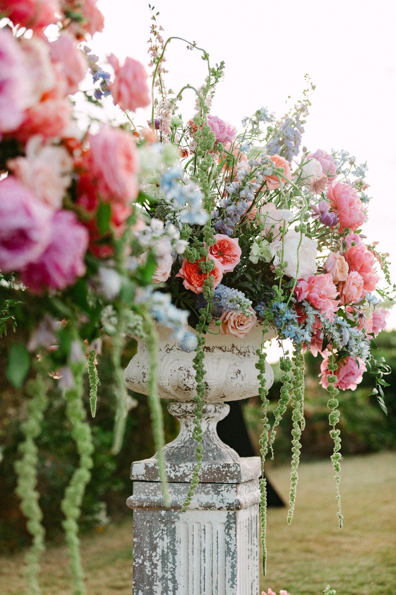 Lush pink, coral, and blue floral arrangement overflows from a vintage stone urn outdoors.
