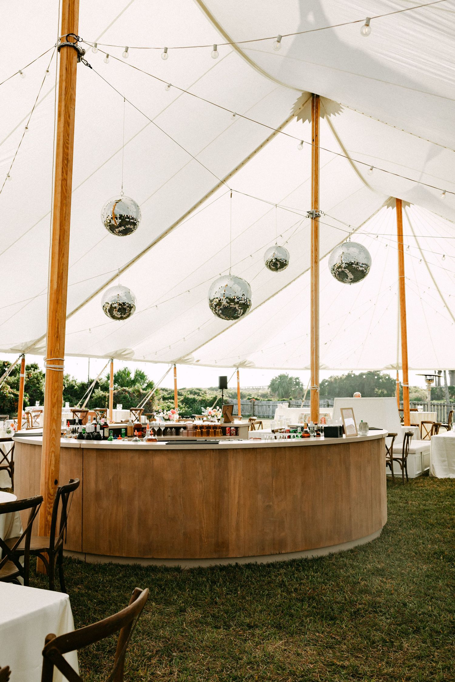Round wooden bar with disco ball decorations under a white sailcloth wedding reception tent.